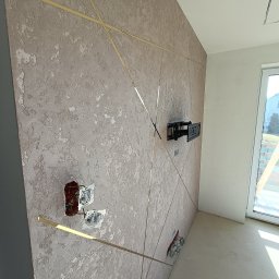 Pro Standard Bartłomiej Miczołek - Teksturowana ściana z dekoracyjnymi, złotymi listwami, uchwyt TV na ścianie, niezakończone gniazdka elektryczne, surowa podłoga, widok na okno z zewnątrz.
