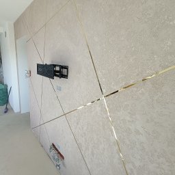 Pro Standard Bartłomiej Miczołek - Teksturowana ściana z dekoracyjnymi, złotymi listwami tworzącymi geometryczny wzór, uchwyt na telewizor zamontowany na ścianie, widoczne kable elektryczne w ścianie, pomieszczenie w trakcie...