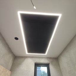 Pro Standard Bartłomiej Miczołek - Czarny sufit z podświetleniem LED w kształcie trapezu, widoczne kable elektryczne, ściany w surowym stanie przed wykończeniem, okno z widokiem na drzewa.