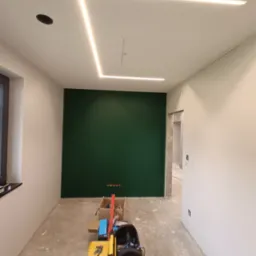 Wnętrze w trakcie wykańczania: Ściana pomalowana na ciemnozielono, sufit z liniowym oświetleniem LED w kształcie prostokąta, widoczne narzędzia i skrzynki narzędziowe na podłodze, przygotowane...