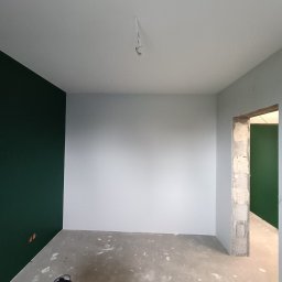 Pro Standard Bartłomiej Miczołek - Wnętrze w trakcie malowania: ściany częściowo w kolorze butelkowej zieleni i szarej bieli, surowy beton na podłodze, widoczny odkurzacz przemysłowy w rogu, niedokończone obramowanie drzwi.