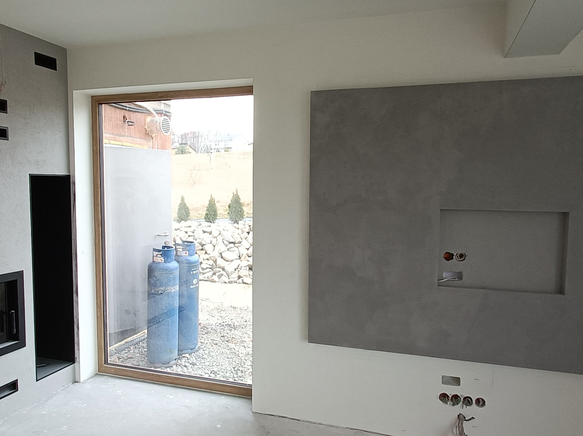 Wnętrze w trakcie wykańczania: ściana z szarą dekoracyjną fakturą, widoczne puszki elektryczne, okno z widokiem na zewnątrz, gdzie stoją dwie niebieskie butle gazowe.