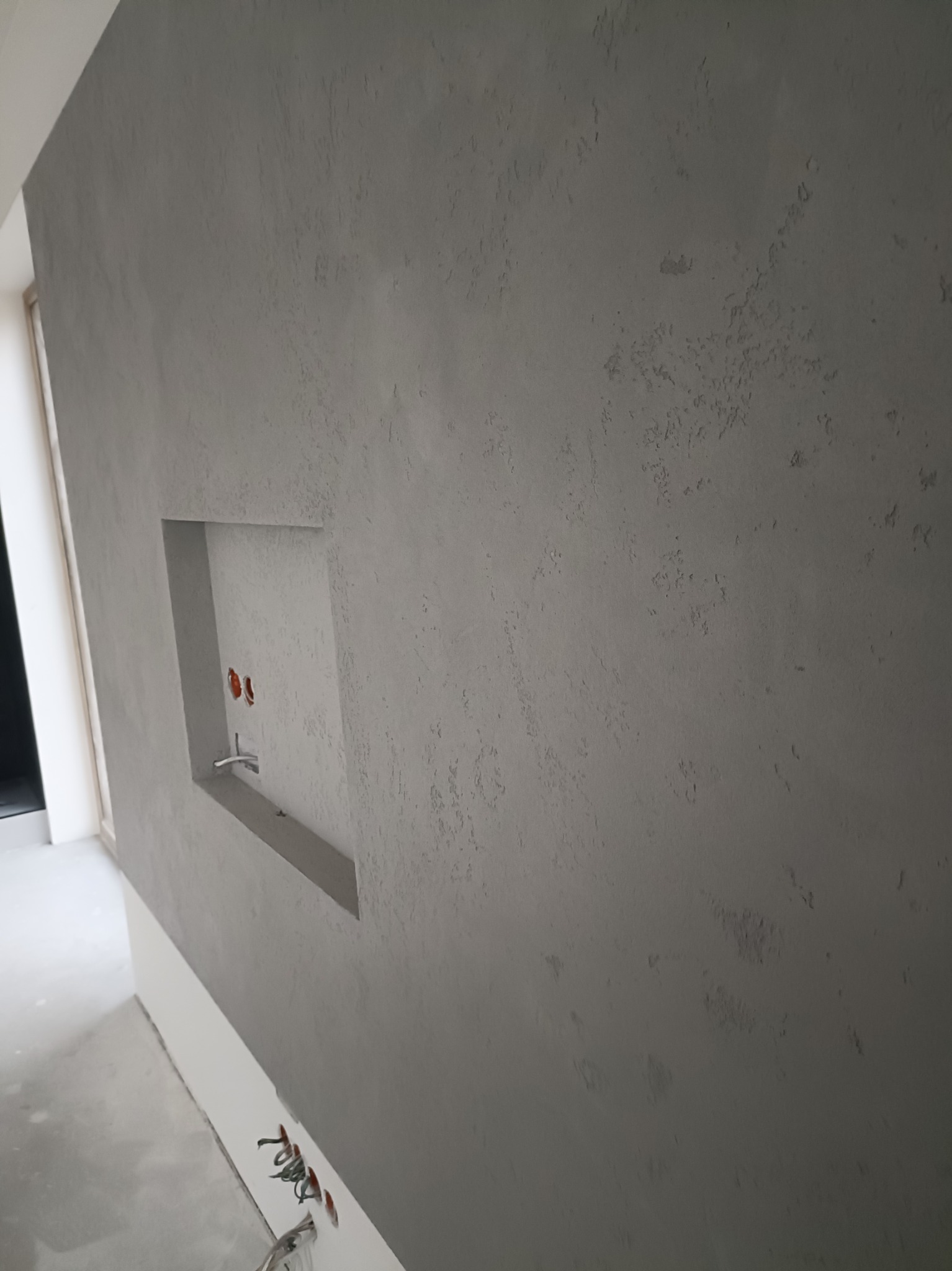 Szara ściana z dekoracyjną strukturą i wnęką z wyprowadzonymi przewodami elektrycznymi w surowym stanie, widoczne kable przy podłodze.