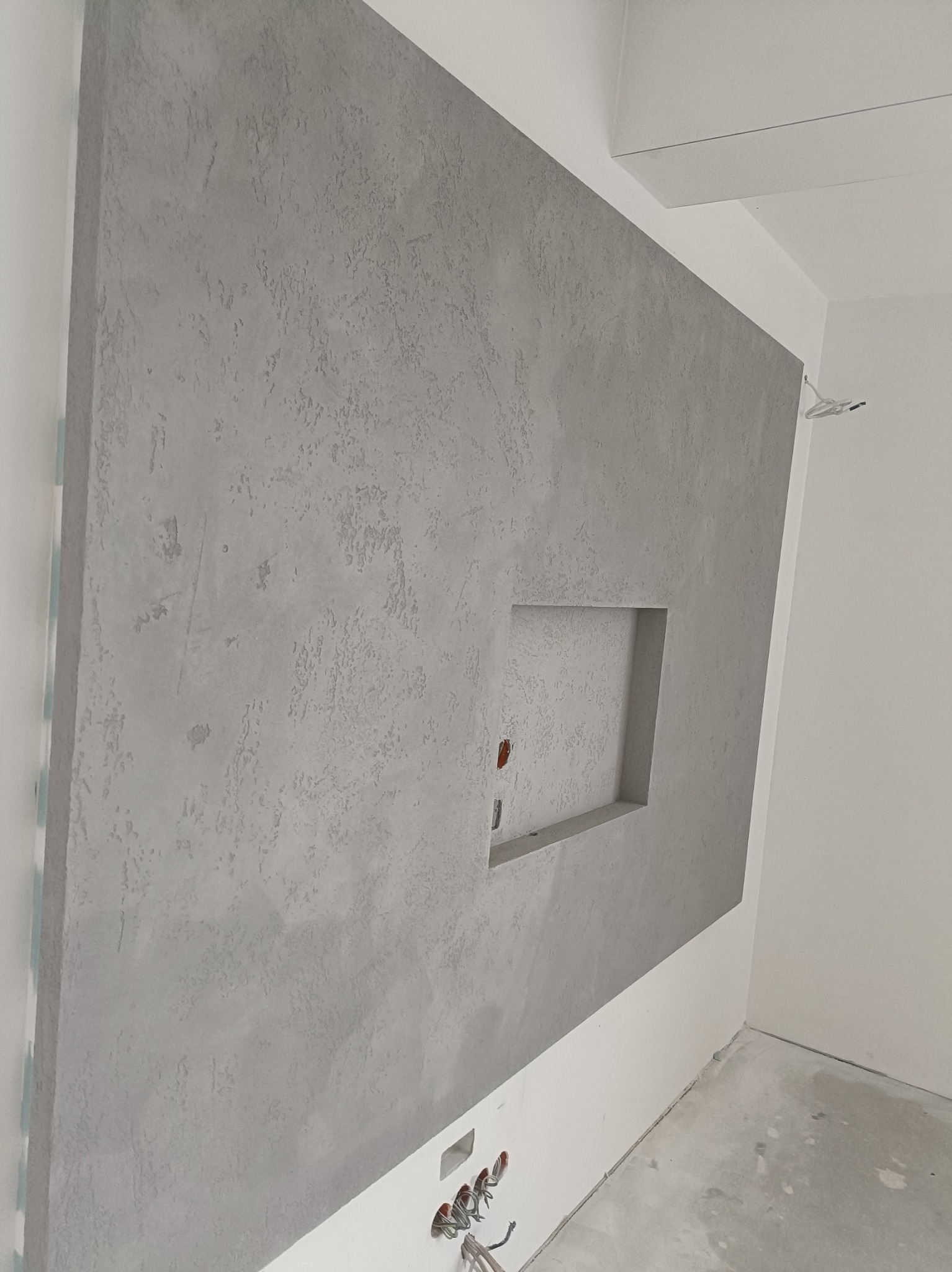 Szara ściana z dekoracyjną fakturą i wnęką, widoczne instalacje elektryczne w dolnej części ściany i wewnątrz wnęki, surowy beton na podłodze.