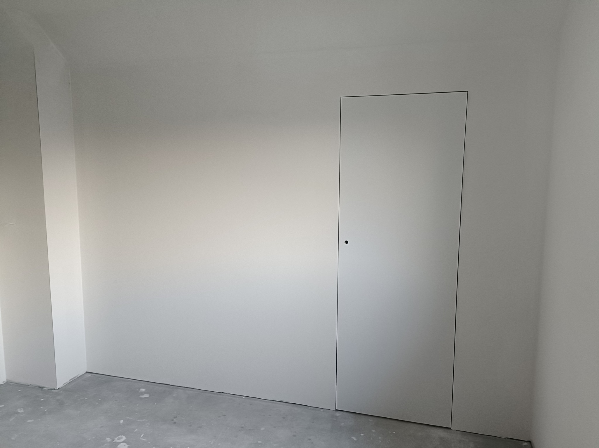 Wnętrze w trakcie prac wykończeniowych, widoczna ściana z ukrytymi drzwiami, surowy beton na podłodze z resztkami zaprawy.