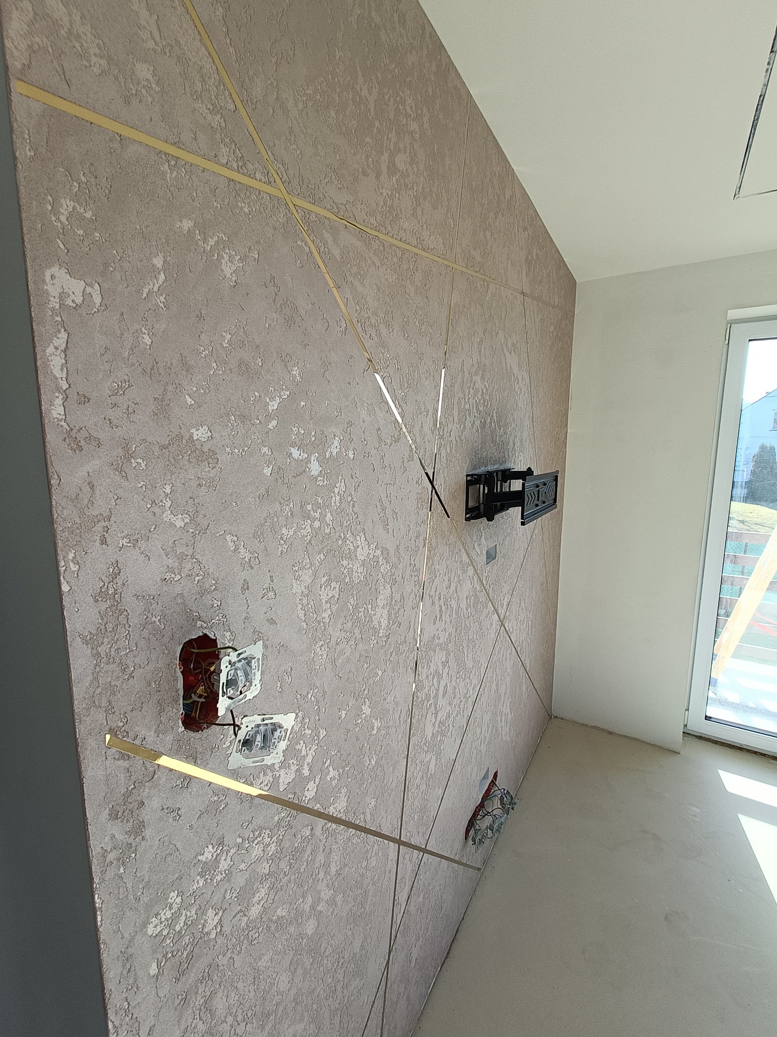 Teksturowana ściana z dekoracyjnymi, złotymi listwami, uchwyt TV na ścianie, niezakończone gniazdka elektryczne, surowa podłoga, widok na okno z zewnątrz.
