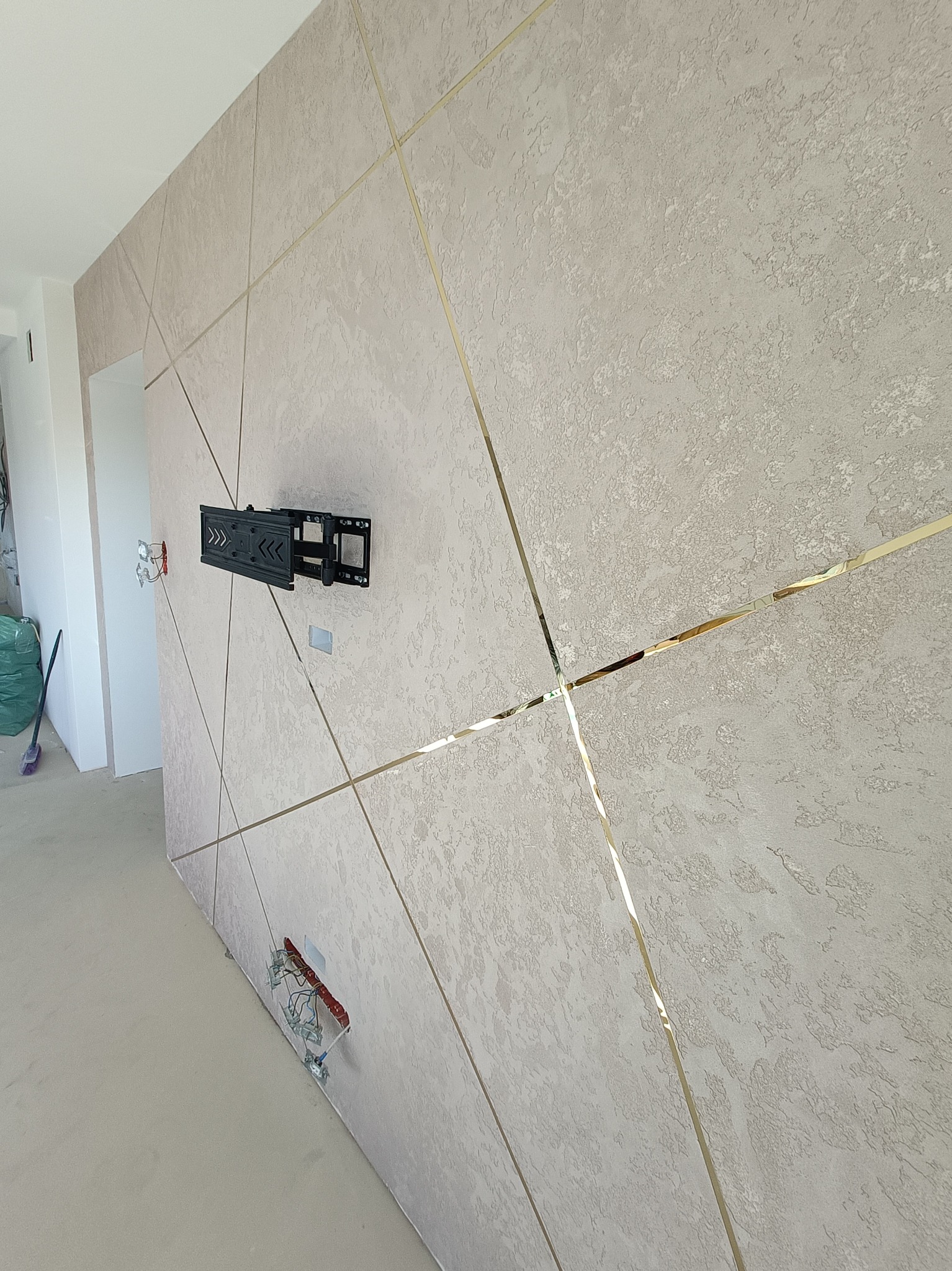 Teksturowana ściana z dekoracyjnymi, złotymi listwami tworzącymi geometryczny wzór, uchwyt na telewizor zamontowany na ścianie, widoczne kable elektryczne w ścianie, pomieszczenie w trakcie...