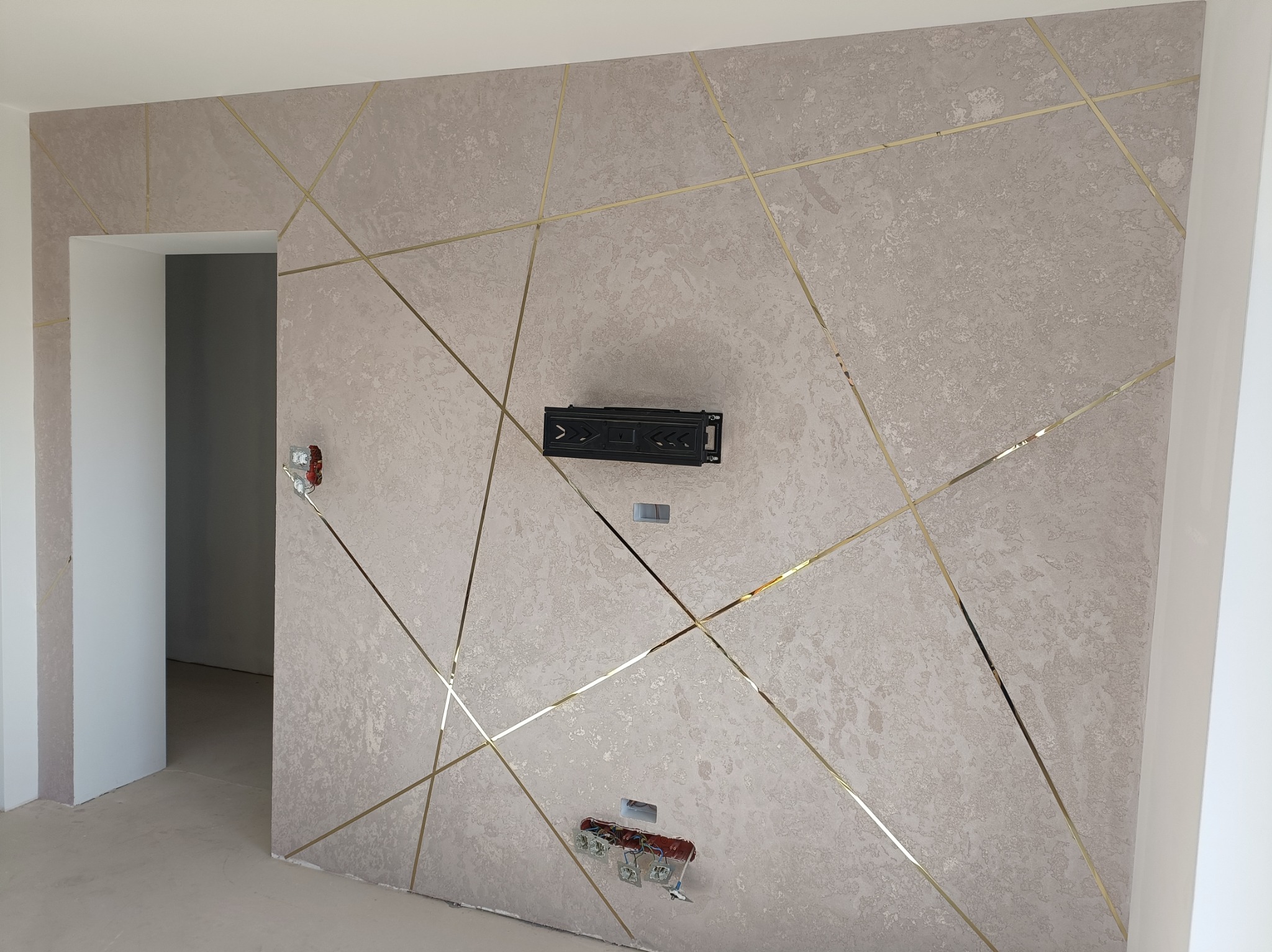 Ściana w pokoju z dekoracyjną strukturą i złotymi listwami tworzącymi geometryczny wzór, widoczne gniazdka elektryczne w trakcie montażu i uchwyt do telewizora.