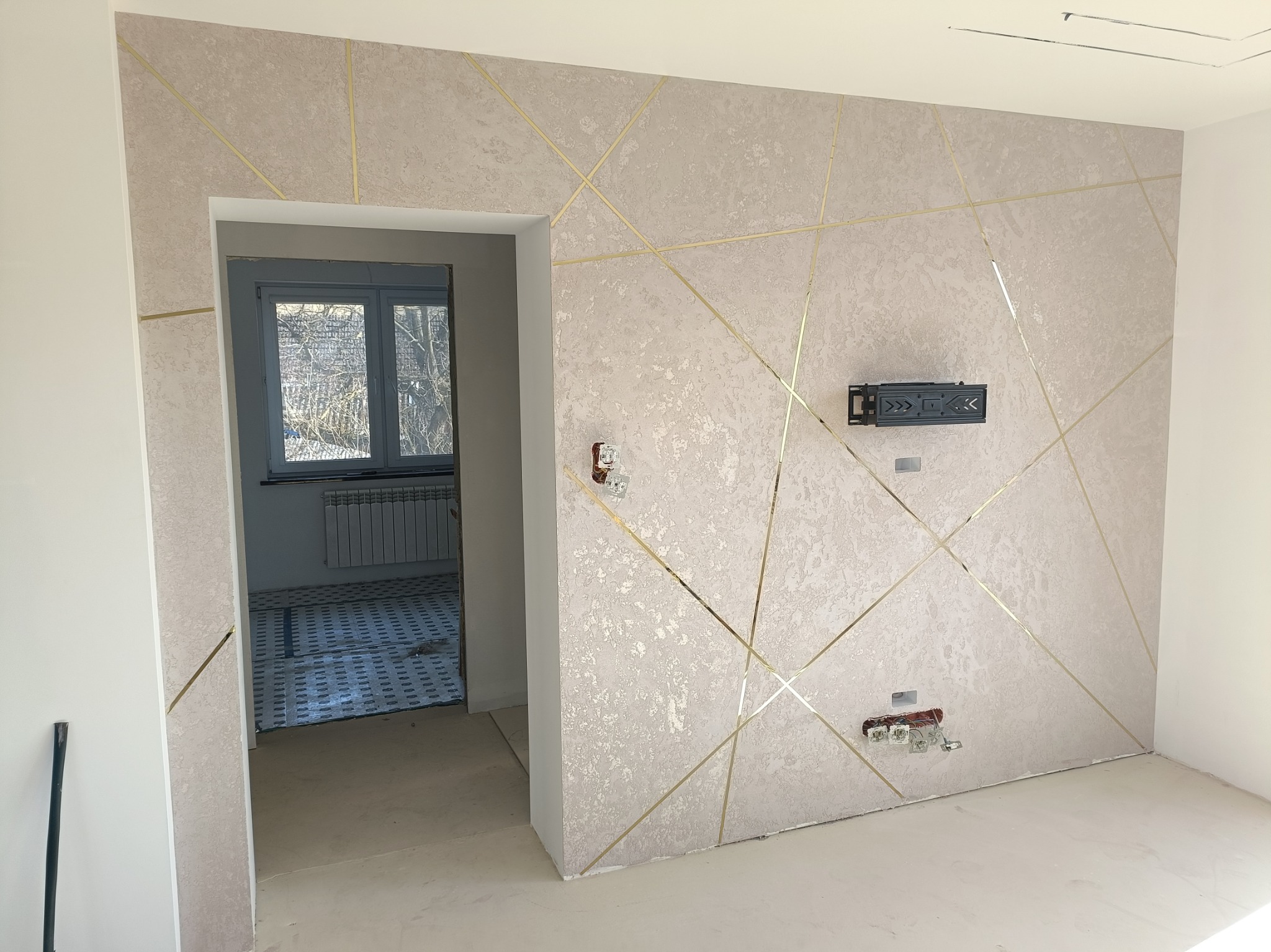 Ściana z dekoracyjną, beżową fakturą i złotymi, ukośnymi listwami, widoczne gniazdka elektryczne i uchwyt na telewizor, w tle widok przez okno z grzejnikiem.