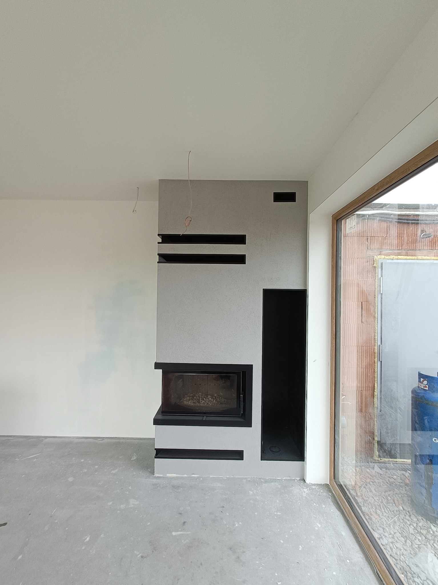 Minimalistyczny kominek z szarego betonu architektonicznego z czarnymi akcentami, w surowym wnętrzu z widocznymi niedokończonymi pracami, dużym oknem i instalacją elektryczną wystającą z sufitu.