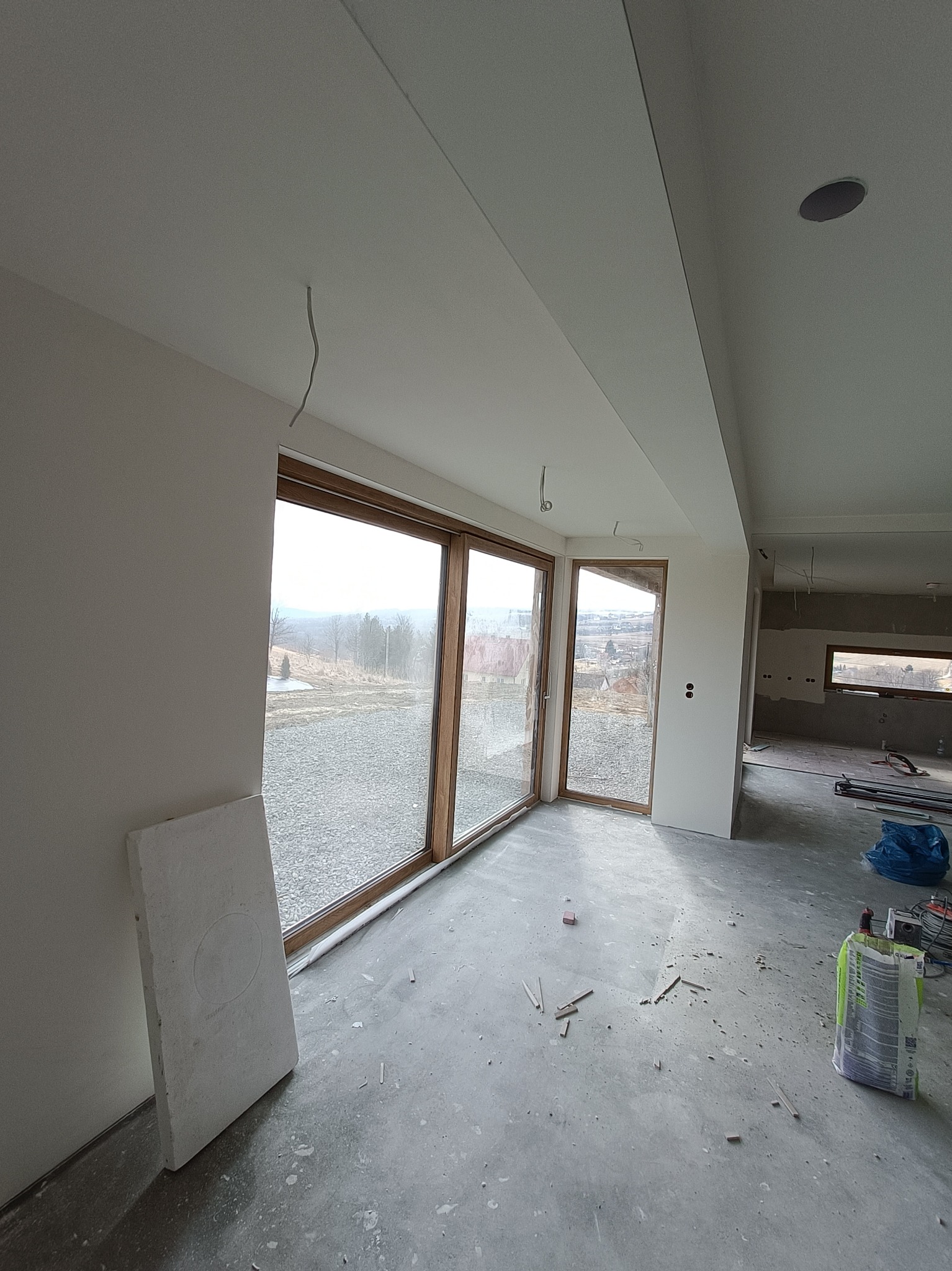 Surowe wnętrze w trakcie prac wykończeniowych: ściany przygotowane pod malowanie, duże okna z widokiem na krajobraz, widoczne kable i materiały budowlane na podłodze.