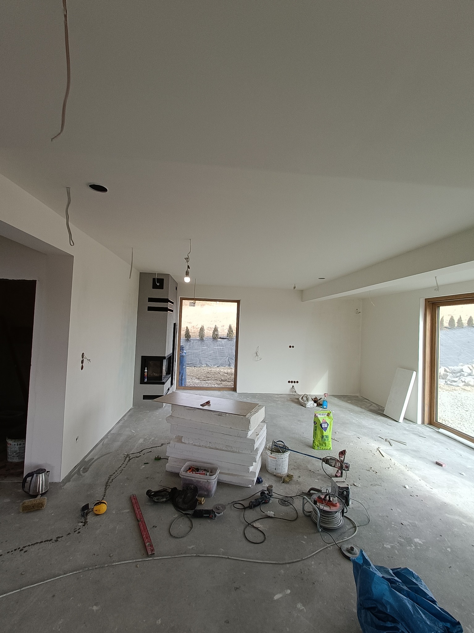 Wnętrze w trakcie prac wykończeniowych: świeżo pomalowane ściany, widoczny kominek z czarnymi akcentami, porozrzucane narzędzia i materiały budowlane na szarym, surowym podłożu, w tle okno...