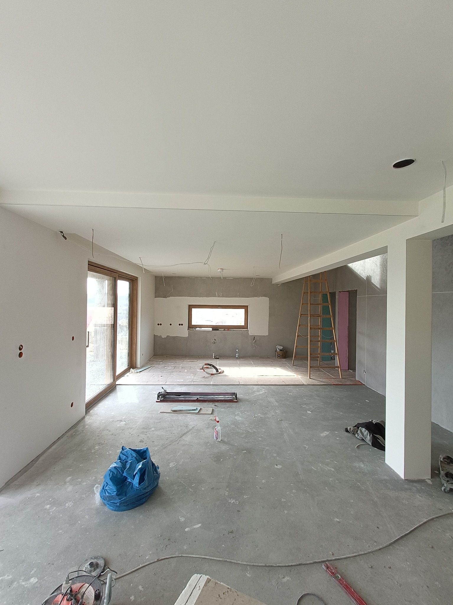 Przestronne wnętrze w trakcie remontu, widoczne ślady po pracach, drabina oparta o ścianę, okno, instalacja elektryczna na suficie, narzędzia i worki budowlane na podłodze.
