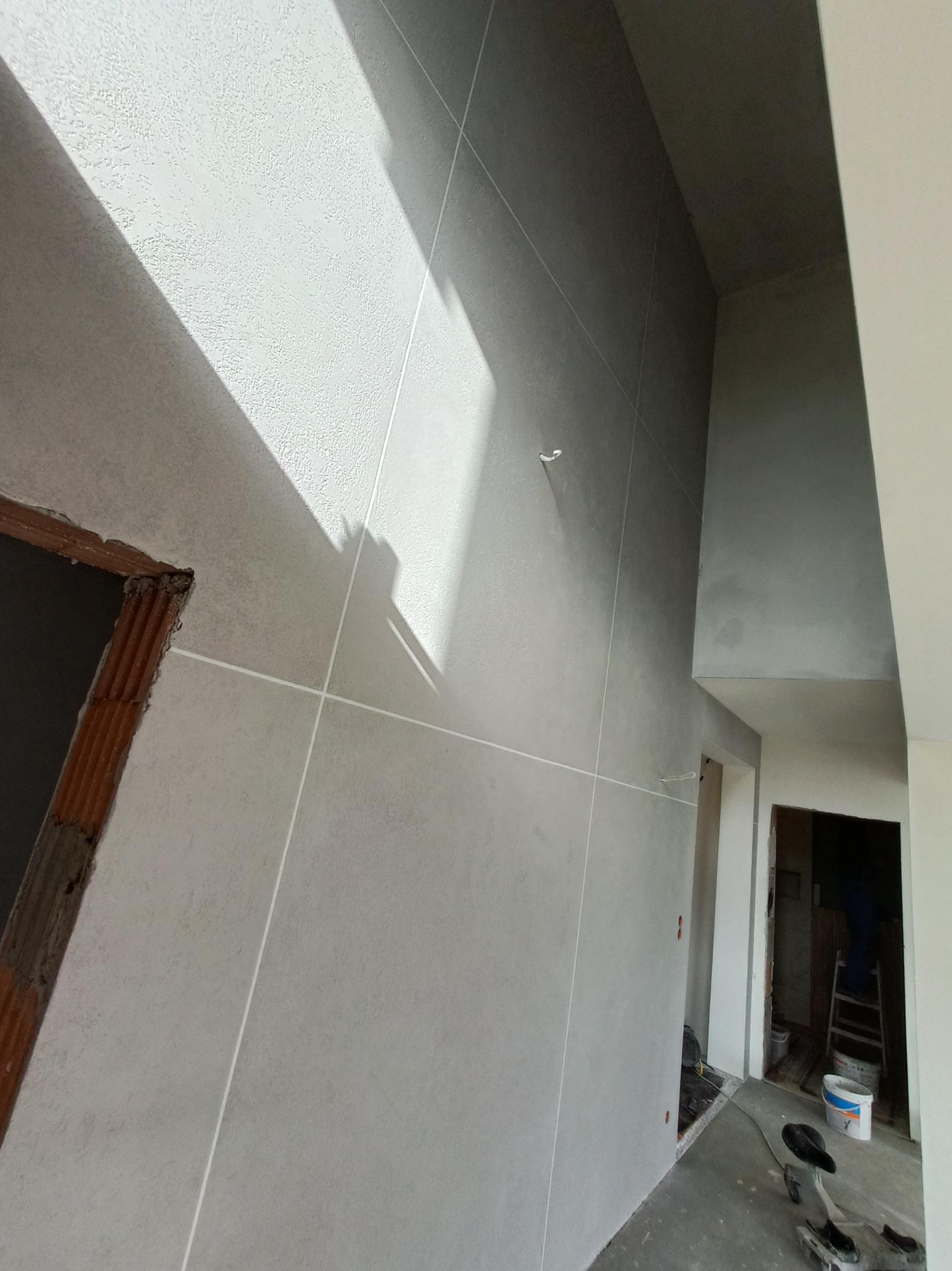 Ściana po nałożeniu gładzi, z widocznymi liniami pionowymi i poziomymi, przygotowana do malowania, z surową ościeżnicą drzwiową po lewej stronie i fragmentem pomieszczenia z drabiną w tle.
