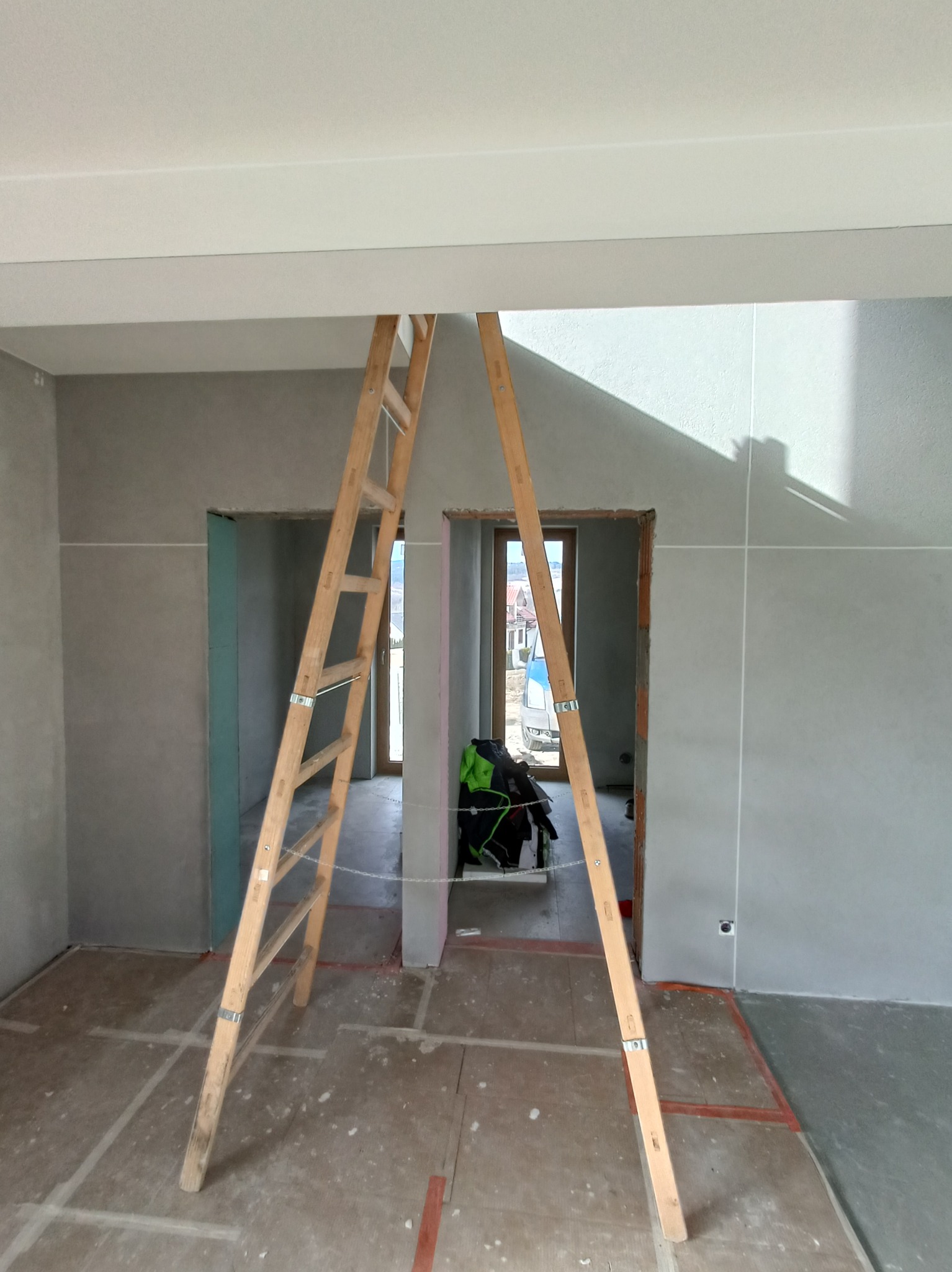 Wnętrze w trakcie remontu: drewniana drabina stoi na podłodze zabezpieczonej folią, ściany pokryte świeżo nałożoną szarą gładzią, widoczne przygotowane otwory drzwiowe.