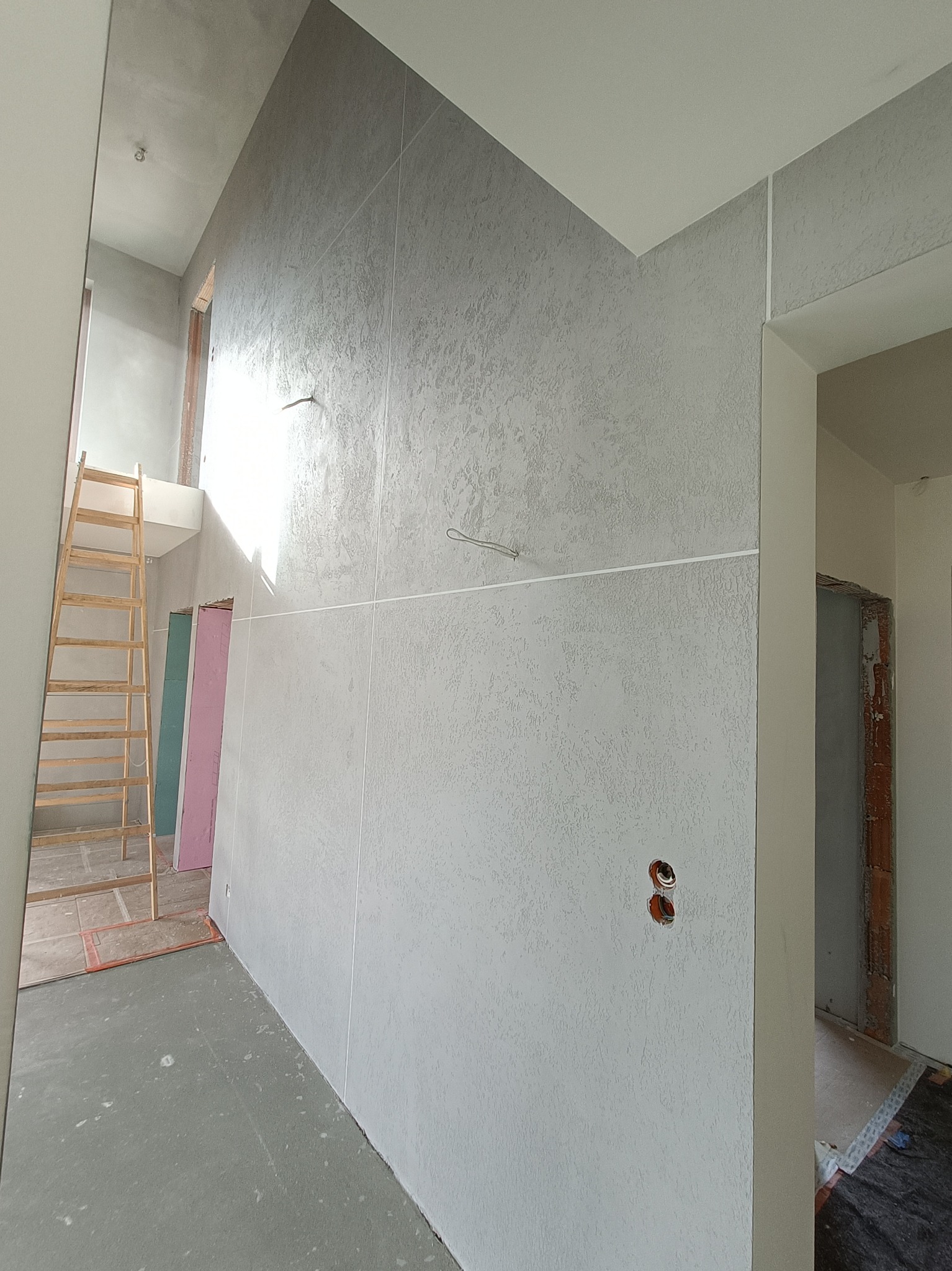 Wnętrze w trakcie prac wykończeniowych: ściany pokryte szarą strukturą, widoczna drabina drewniana, wystające przewody elektryczne i surowe ościeżnice drzwiowe.