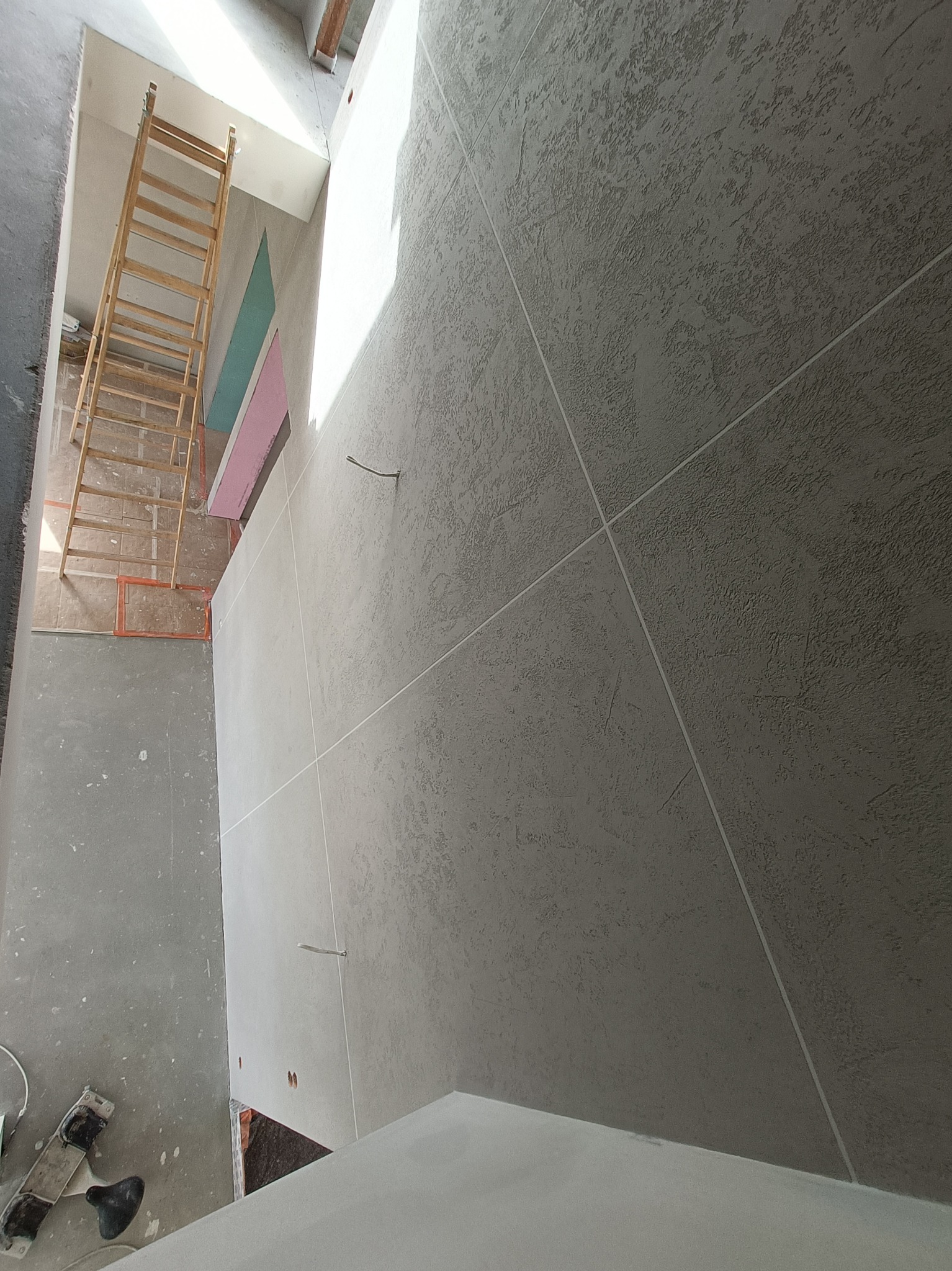 Ściana z szarą, strukturalną gładzią, podzielona na kwadraty, w tle drabina i fragment ocieplonego poddasza z widoczną wełną mineralną i folią w kolorach różowym i zielonym.