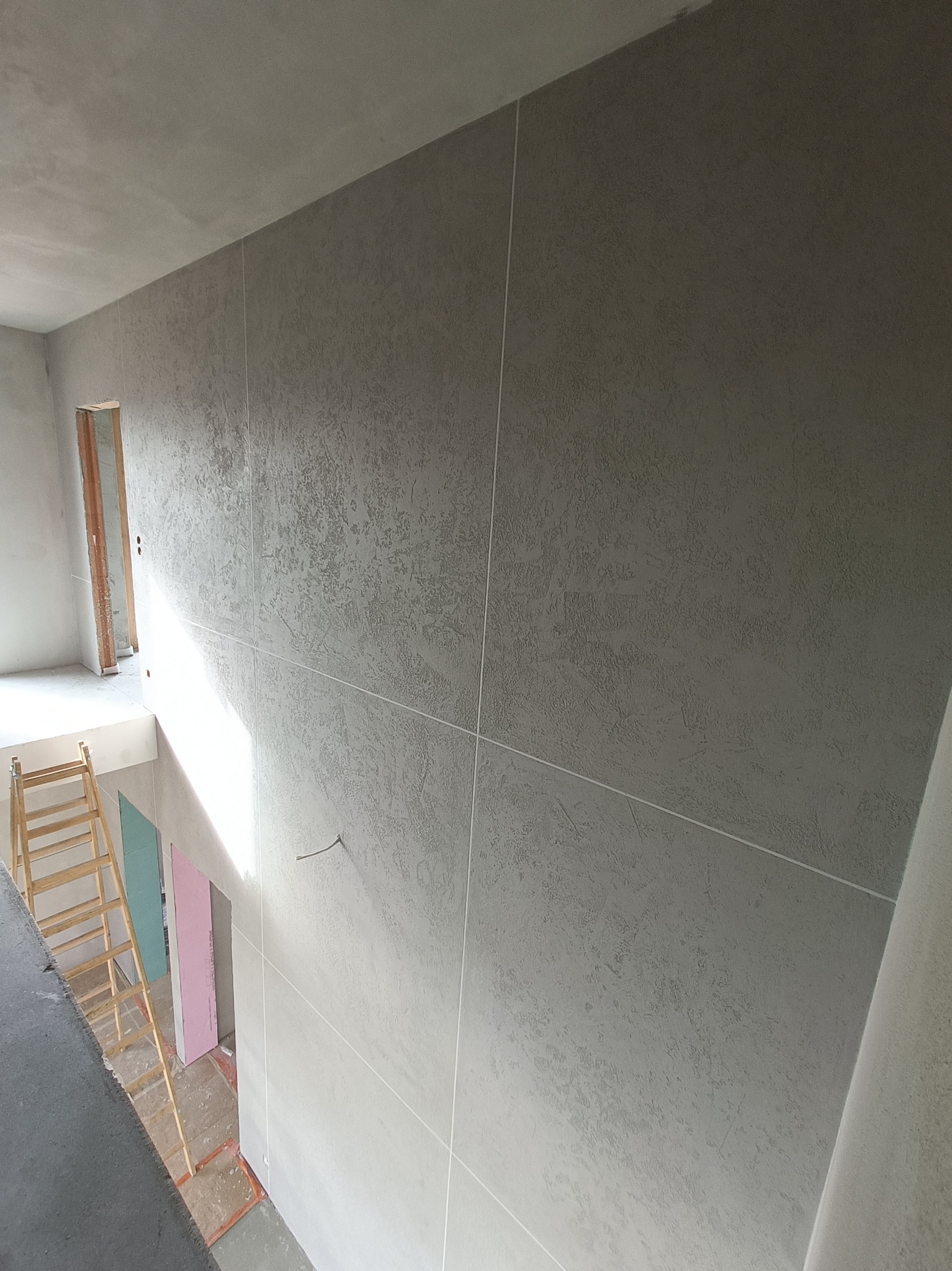 Ściana z nałożoną szarą, dekoracyjną strukturą, podzielona pionowymi i poziomymi liniami, widoczny fragment drabiny i otwór drzwiowy w tle.