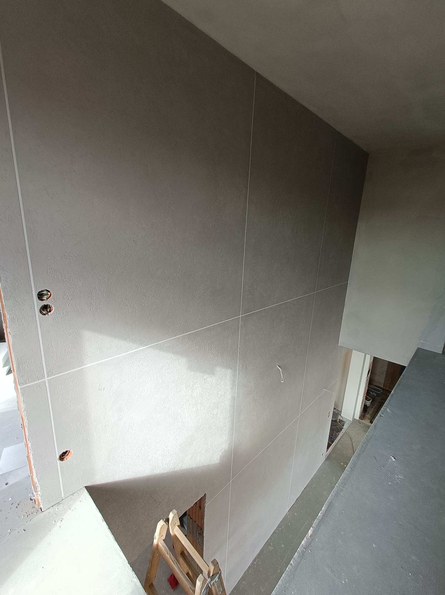 Wnętrze w trakcie prac wykończeniowych, widok z góry na ścianę z wygładzoną powierzchnią i widocznymi puszkami elektrycznymi, fragment drabiny malarskiej w rogu.