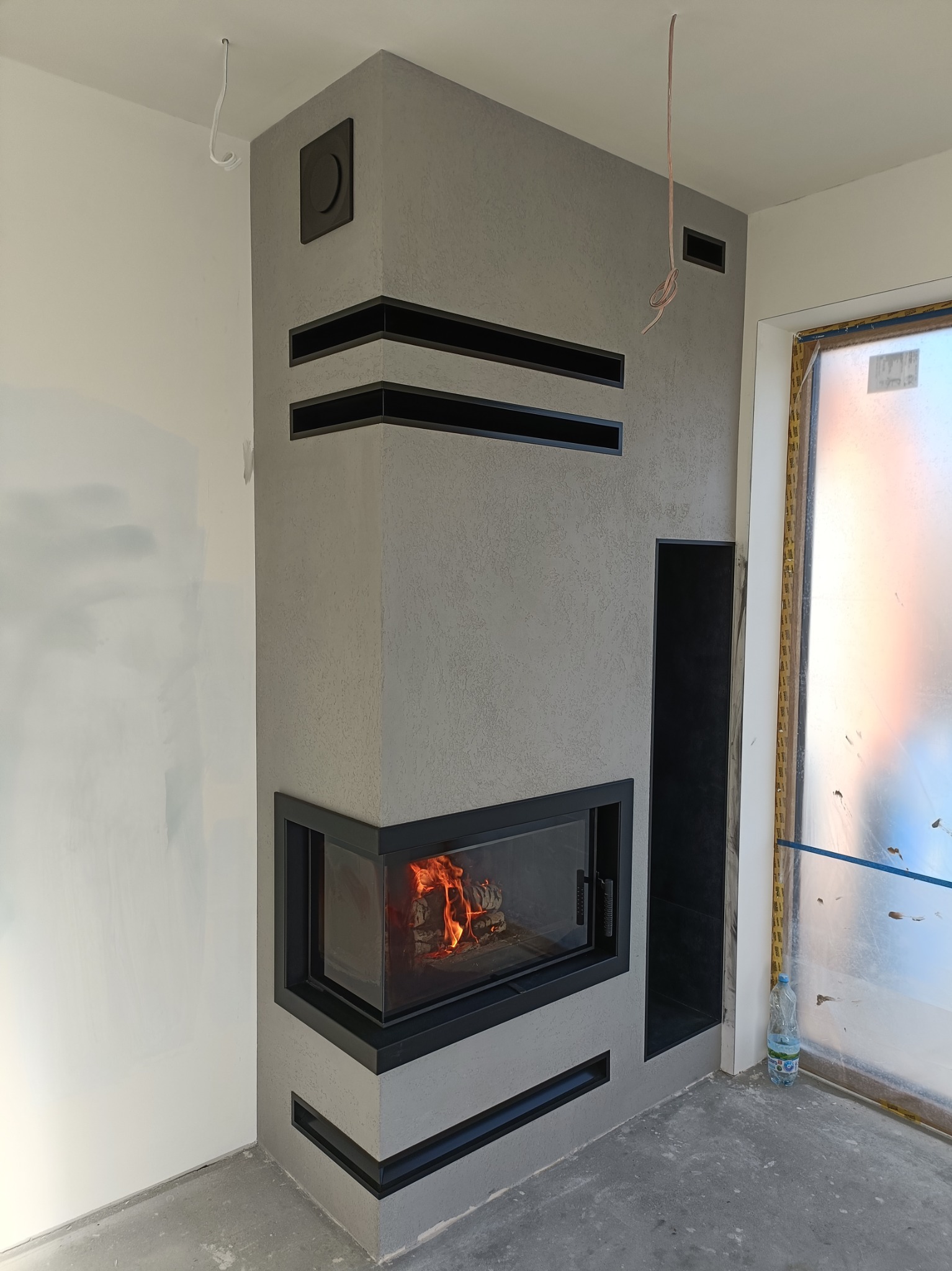 Narożny kominek obudowany szarym tynkiem strukturalnym z czarnymi wstawkami, płomienie widoczne za szybą, surowy stan wykończenia pomieszczenia z widocznymi przewodami i folią ochronną na oknie.
