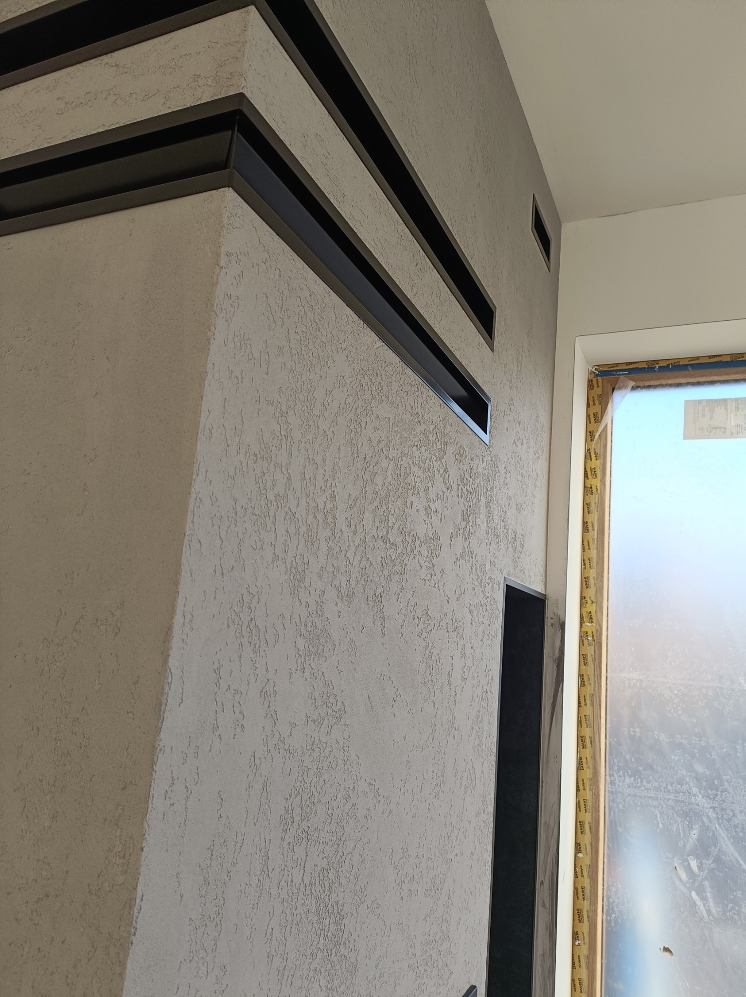 Ściana z dekoracyjną strukturą tynku w odcieniach szarości, z wkomponowanymi czarnymi, poziomymi elementami dekoracyjnymi, widoczna w pobliżu okna zabezpieczonego folią.