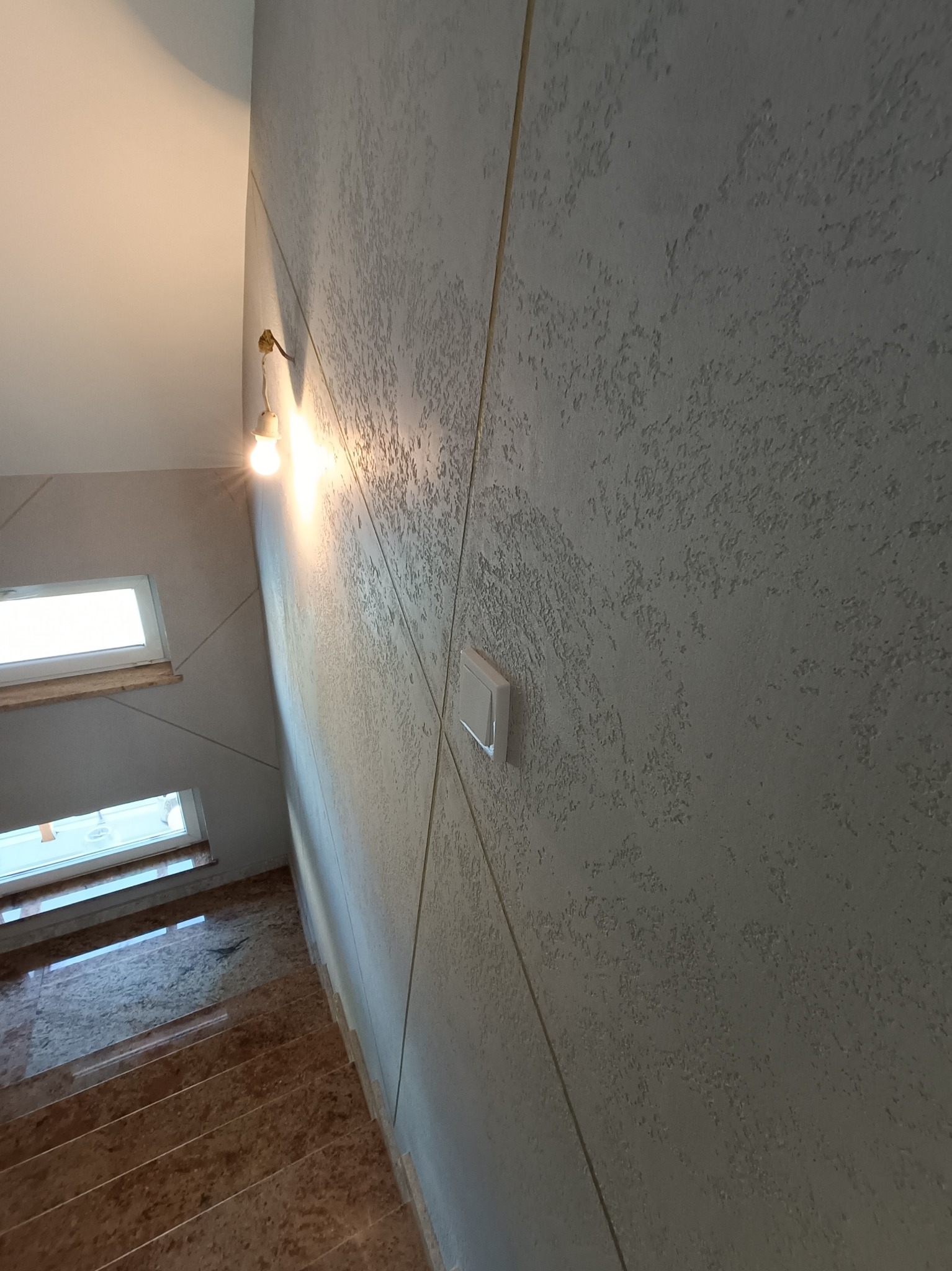 Klatka schodowa z dekoracyjną szarą ścianą o wyraźnej fakturze, oświetlona żarówką, z widocznymi marmurowymi schodami i oknami.