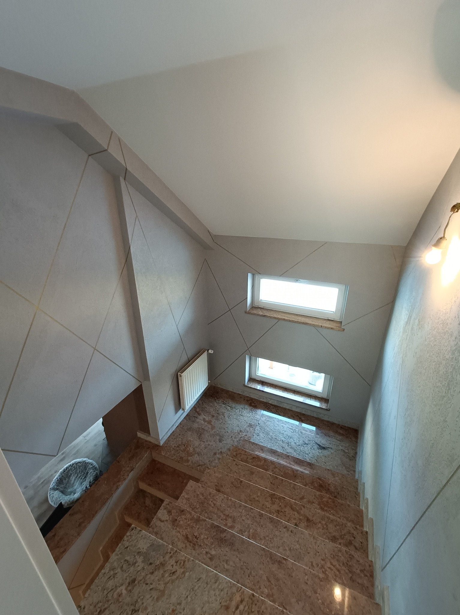 Klatka schodowa z wykończonymi ścianami w jasnych odcieniach szarości z geometrycznym wzorem, marmurowe schody, dwa małe okna doświetlające przestrzeń, grzejnik na ścianie.