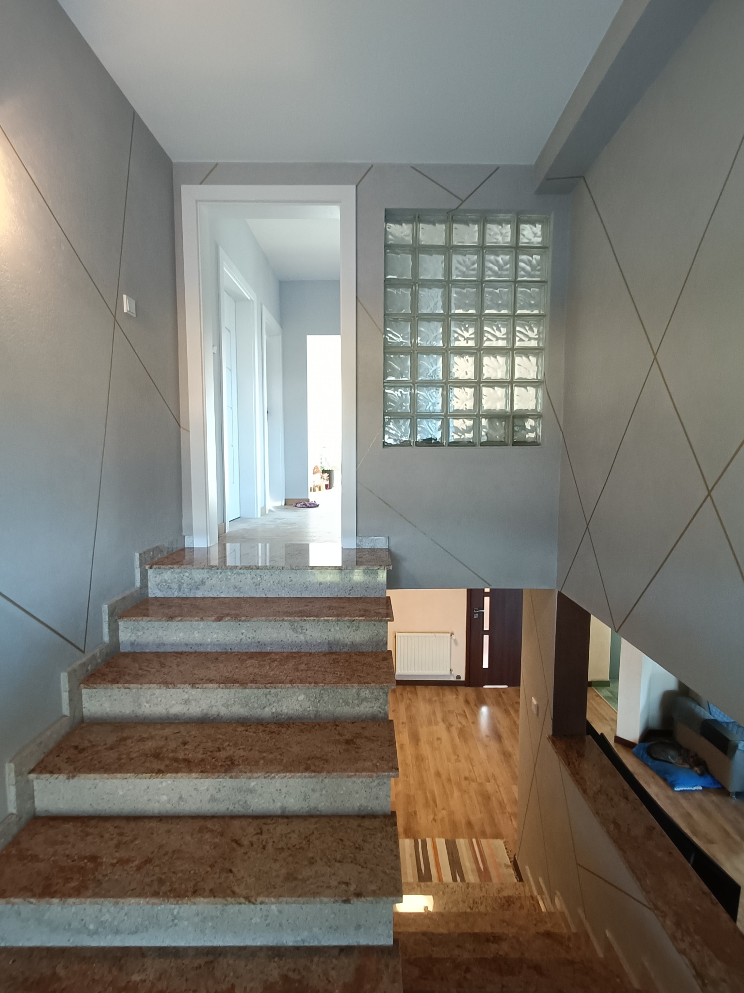 Klatka schodowa z marmurowymi schodami, ściany w odcieniach szarości z geometrycznym wzorem, okno z luksferów, widok na korytarz i salon