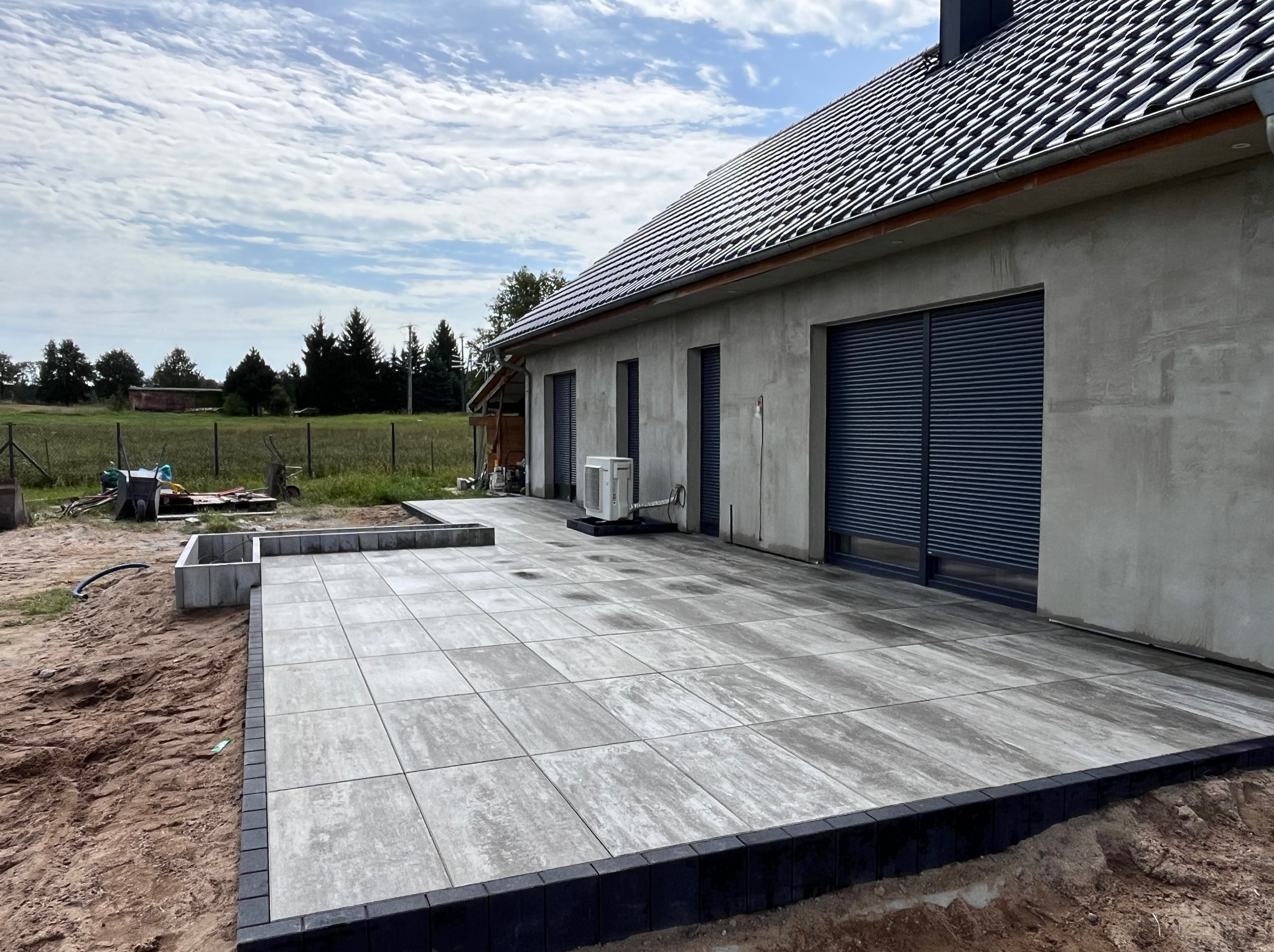 Nowoczesny taras z szarych płyt betonowych obramowany czarną kostką, przylegający do surowego budynku z szarymi roletami okiennymi, widoczne prace wykończeniowe wokół