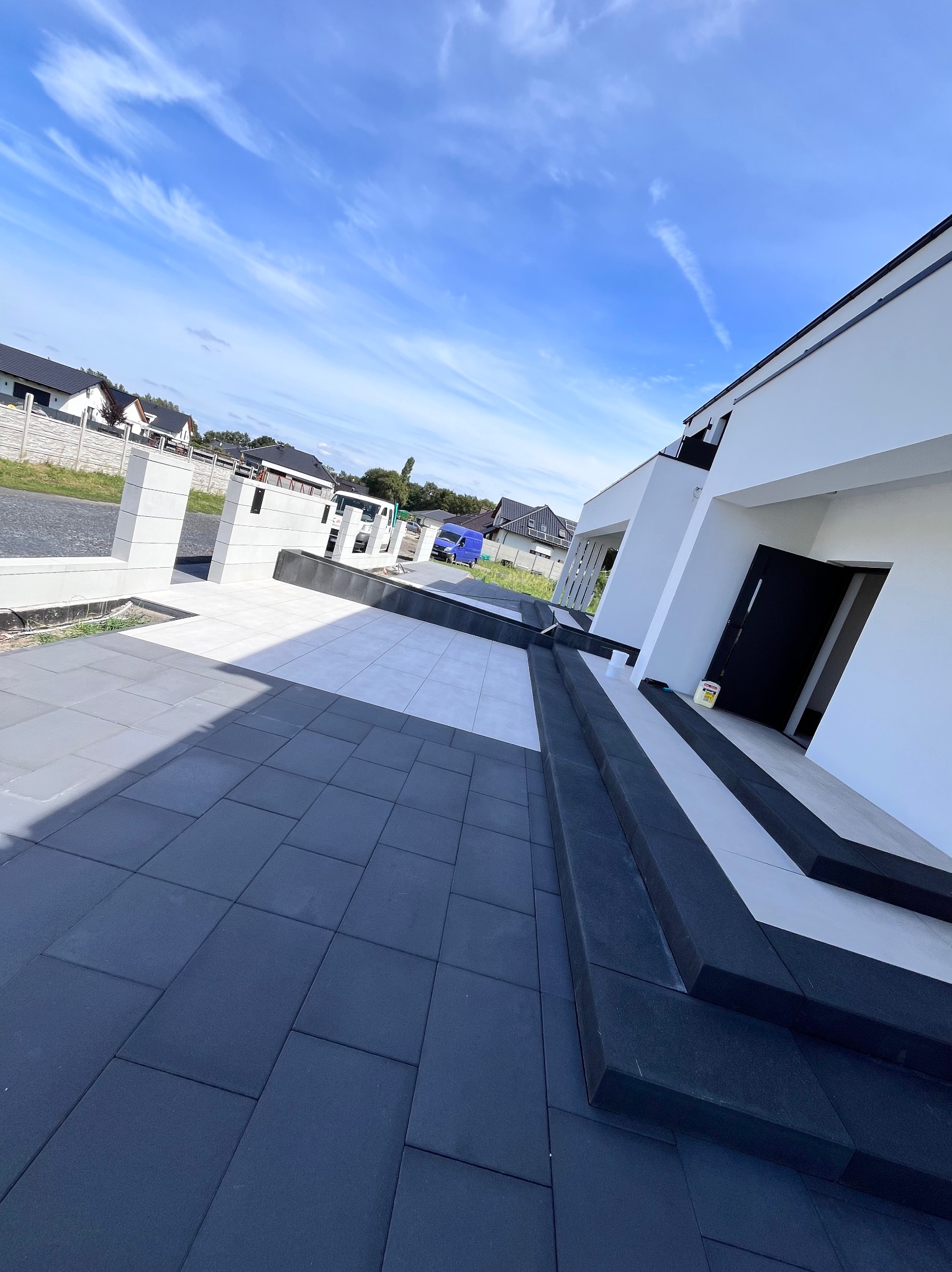 Nowoczesny podjazd wyłożony szarą kostką brukową prowadzący do białego domu z czarnymi akcentami, widoczne schody i elementy dekoracyjne, błękitne niebo w tle.