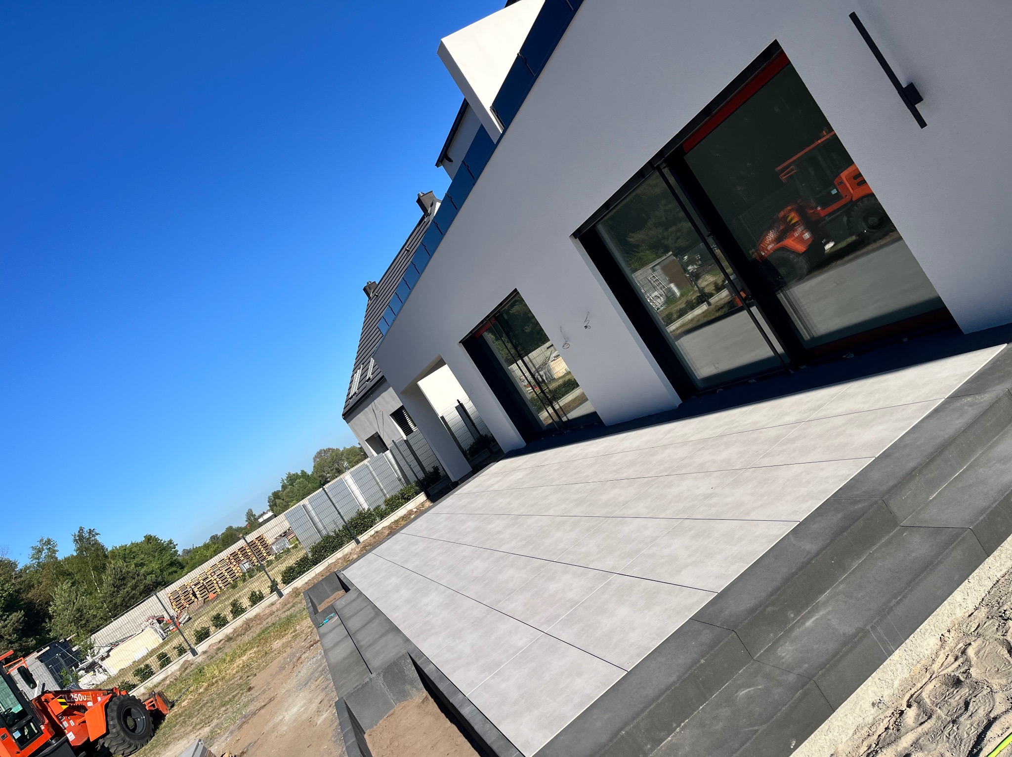 Nowoczesny taras wyłożony jasnoszarą płytką, z widocznym obrzeżem i orange miniładowarką w tle oraz odbiciem większej maszyny w oknie domu.