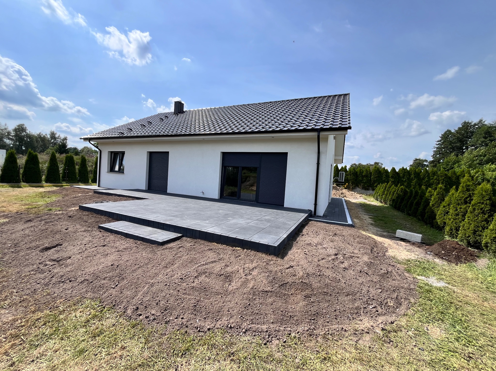 Nowoczesny taras z szarych płyt brukowych w Płotach, z niskimi schodami i widokiem na biały dom z ciemną dachówką i roletami.