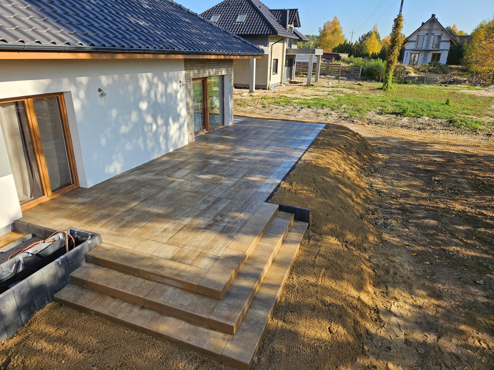 Nowo ułożony taras z kostki brukowej z trzema stopniami prowadzącymi do domu; widoczna niezabezpieczona instalacja pod tarasem, świeżo nawieziona ziemia wokół.