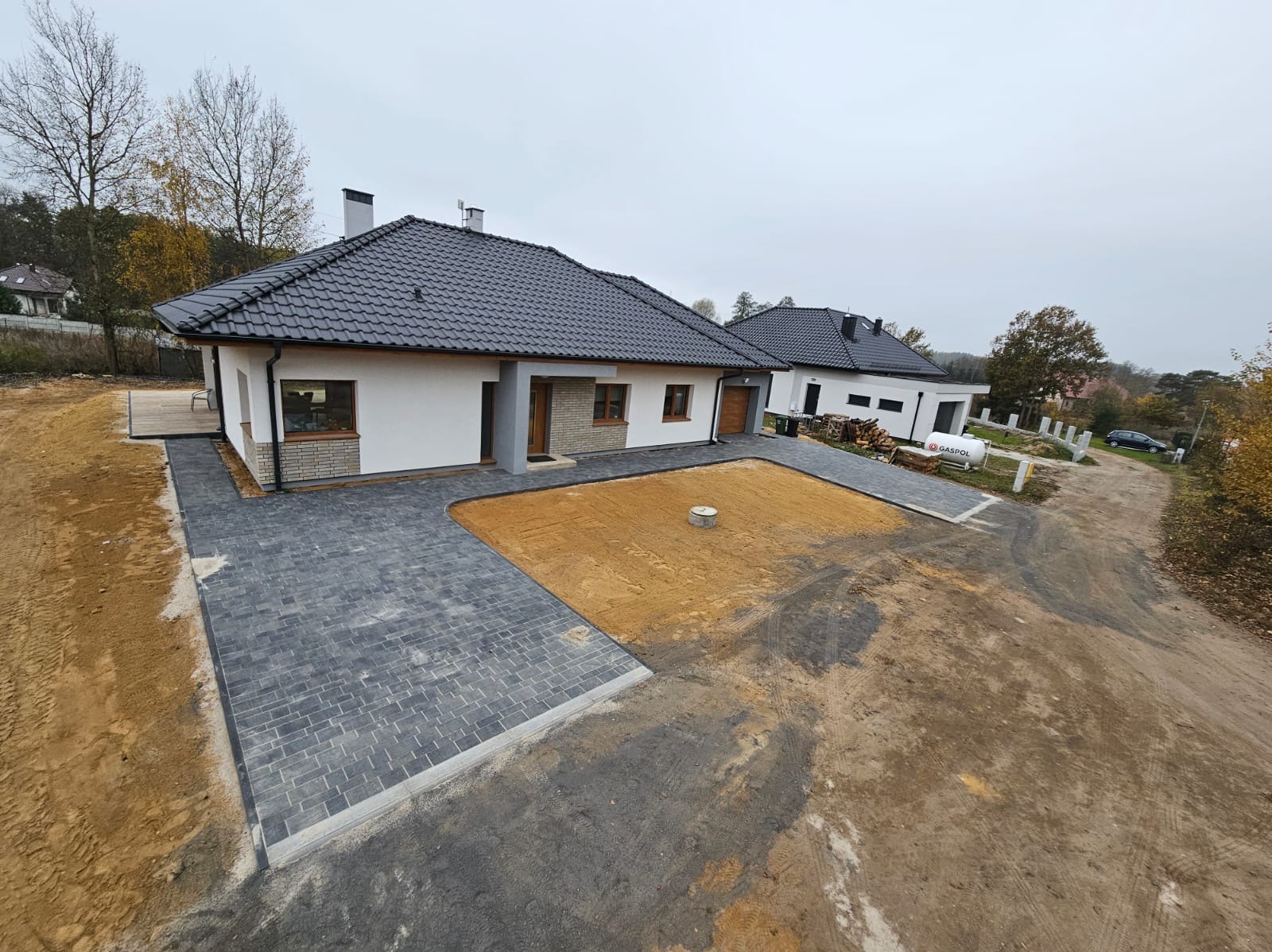 Nowoczesny dom jednorodzinny z szarą kostką brukową ułożoną na podjeździe i ścieżkach, przygotowany teren pod trawnik, widok z góry na posesję w pochmurny dzień.