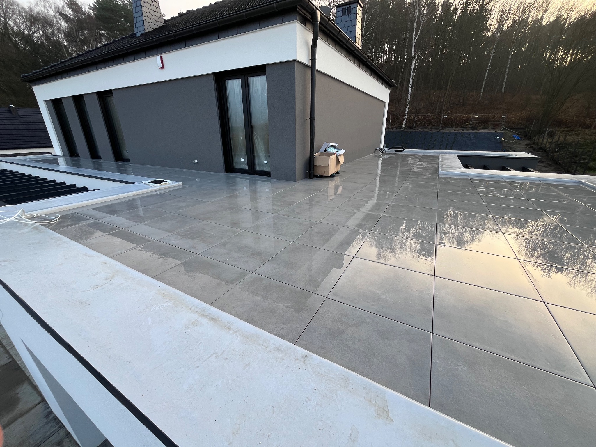 Szare płytki tarasowe ułożone na zewnątrz nowoczesnego domu z widokiem na drzewa. Częściowo widoczny biały murek i kartony z akcesoriami.