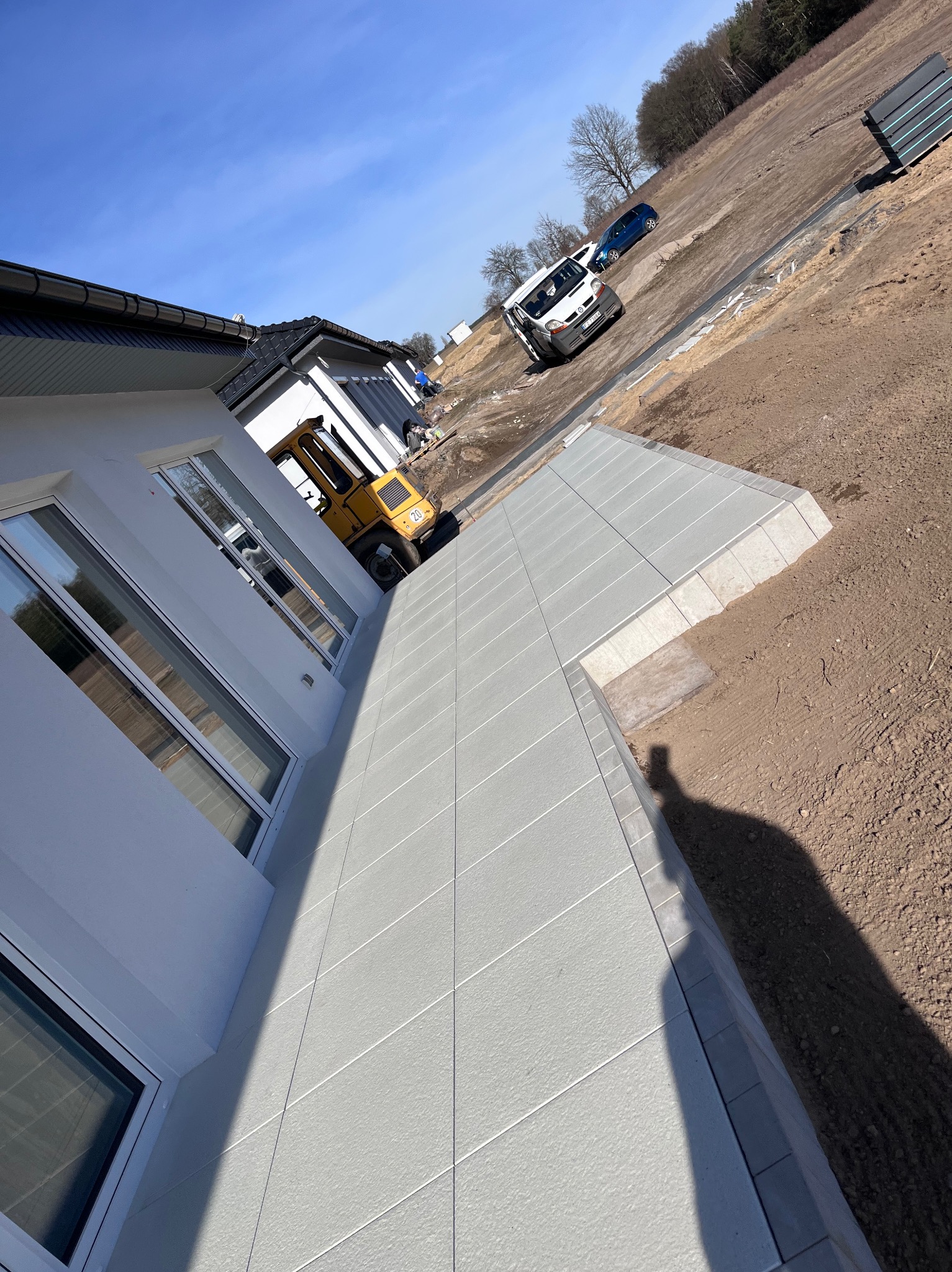 Nowoczesny taras z jasnoszarej kostki, widok z góry, cień osoby, w tle budynek i maszyna budowlana. Słoneczny dzień, błękitne niebo.