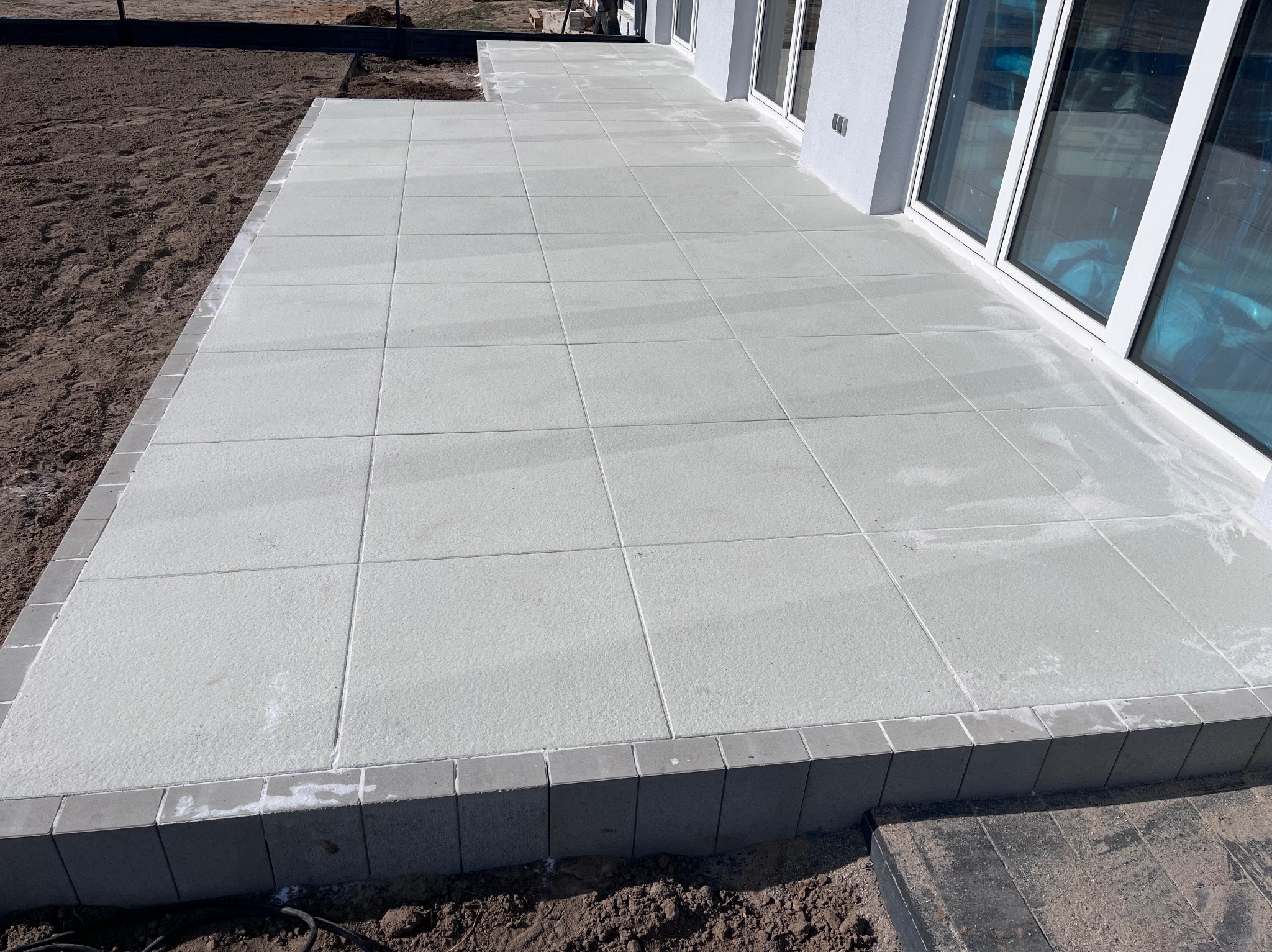 Jasnoszara, kwadratowa kostka brukowa tworząca taras przy budynku z dużymi oknami, obrzeża wykończone ciemniejszą kostką. Częściowo widoczna ziemia.