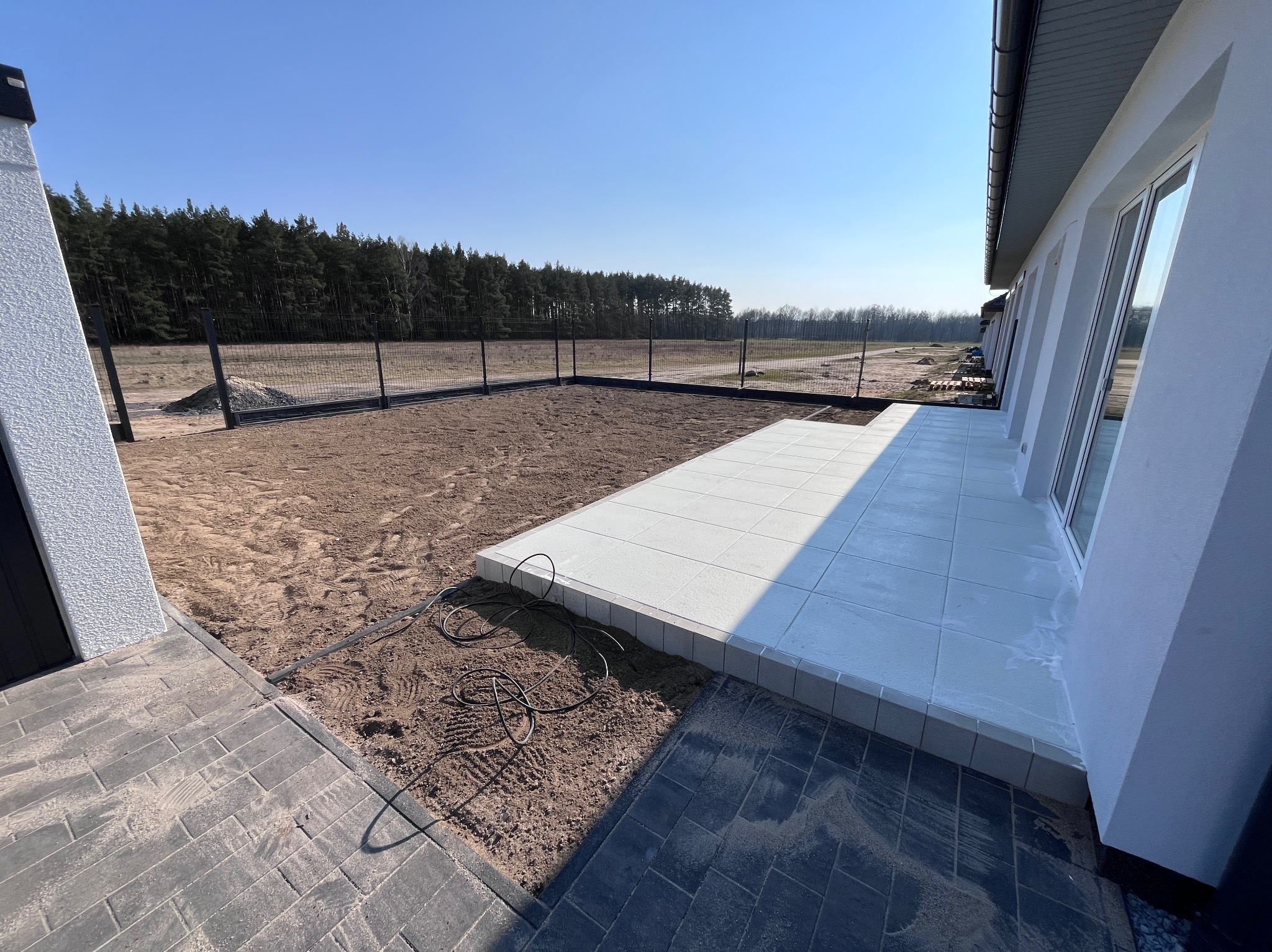 Biała, kwadratowa nawierzchnia tarasu przylegająca do budynku, z widocznym przygotowanym terenem pod dalsze prace i fragmentem ułożonej kostki brukowej.