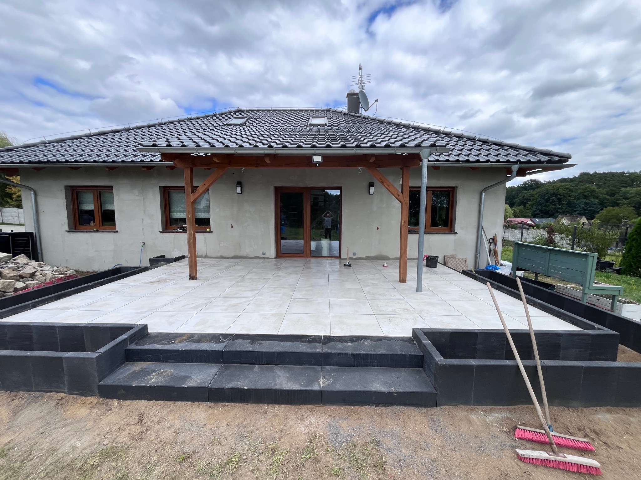 Wykończony taras z jasnych płyt, otoczony czarnymi obrzeżami i schodami, z drewnianą pergolą na tle domu jednorodzinnego i pochmurnego nieba.