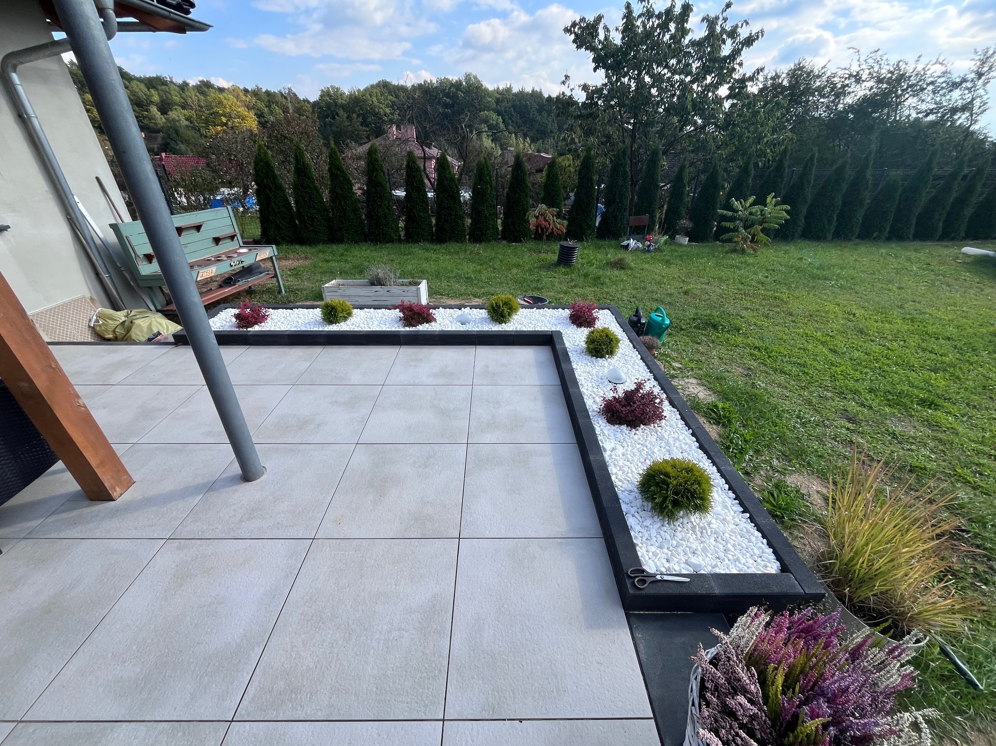 Wykończony taras z jasnych płyt, obrzeża z czarnego kamienia i białych kamyków, ozdobne krzewy, trawa i tuje w tle. Widok na ogród.