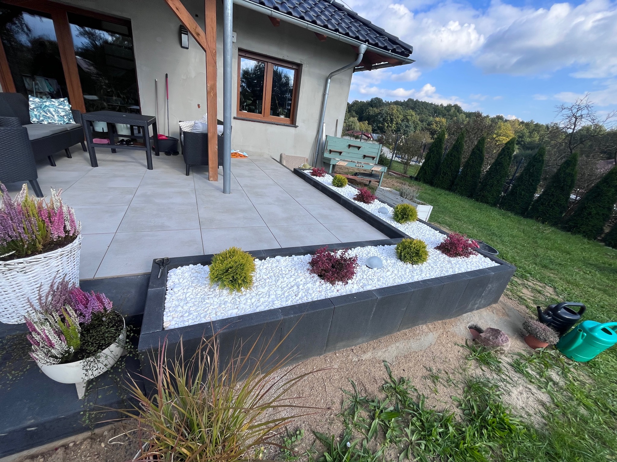 Nowoczesny taras z szarych płyt, obrzeża z kostki granitowej wypełnione białym grysem i ozdobnymi roślinami. W tle dom z drewnianą stolarką okienną.