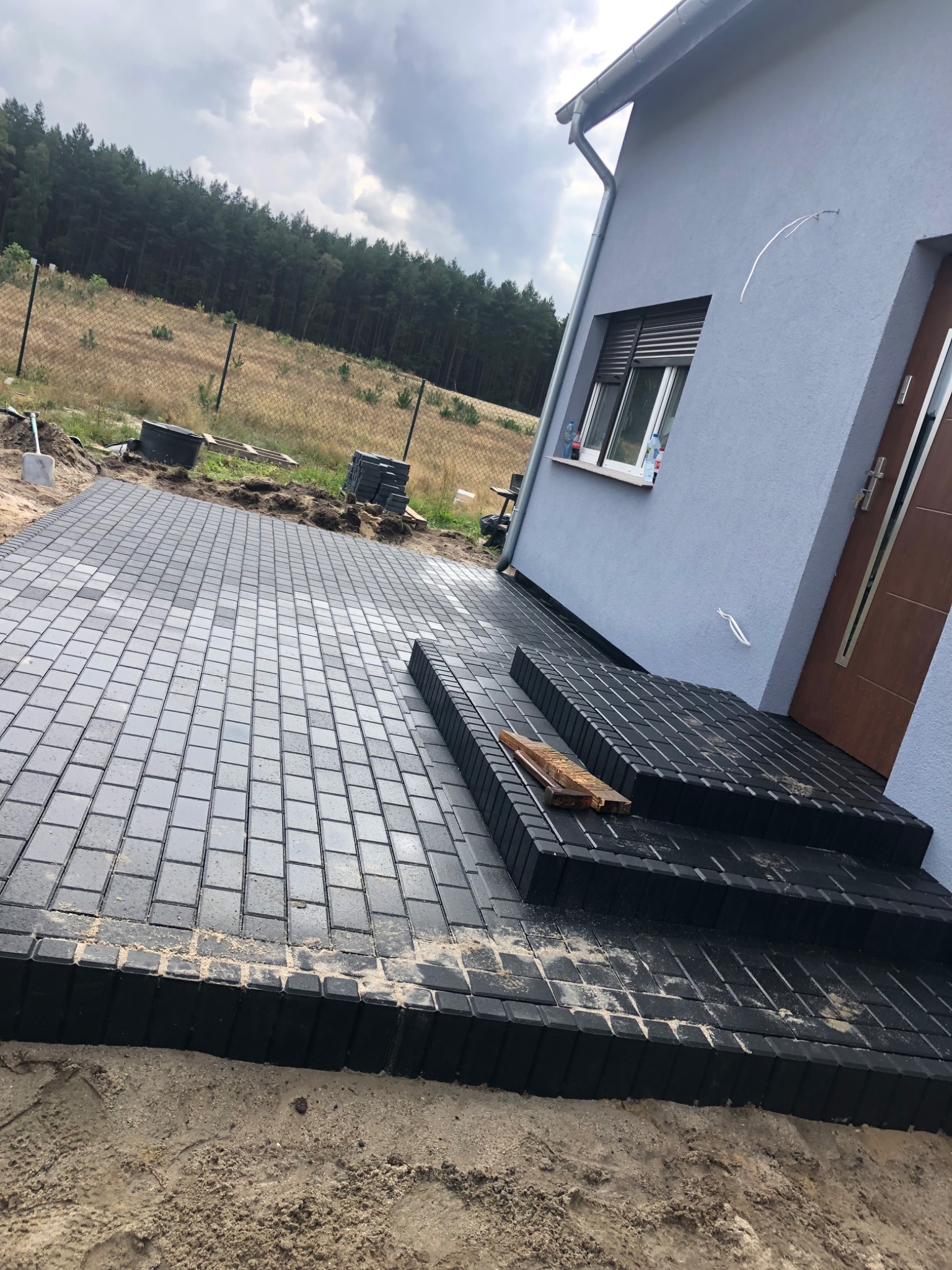 Nowo ułożona, ciemnoszara kostka brukowa przed domem z trzema schodami, widoczne narzędzia i zapas kostki, w tle las.