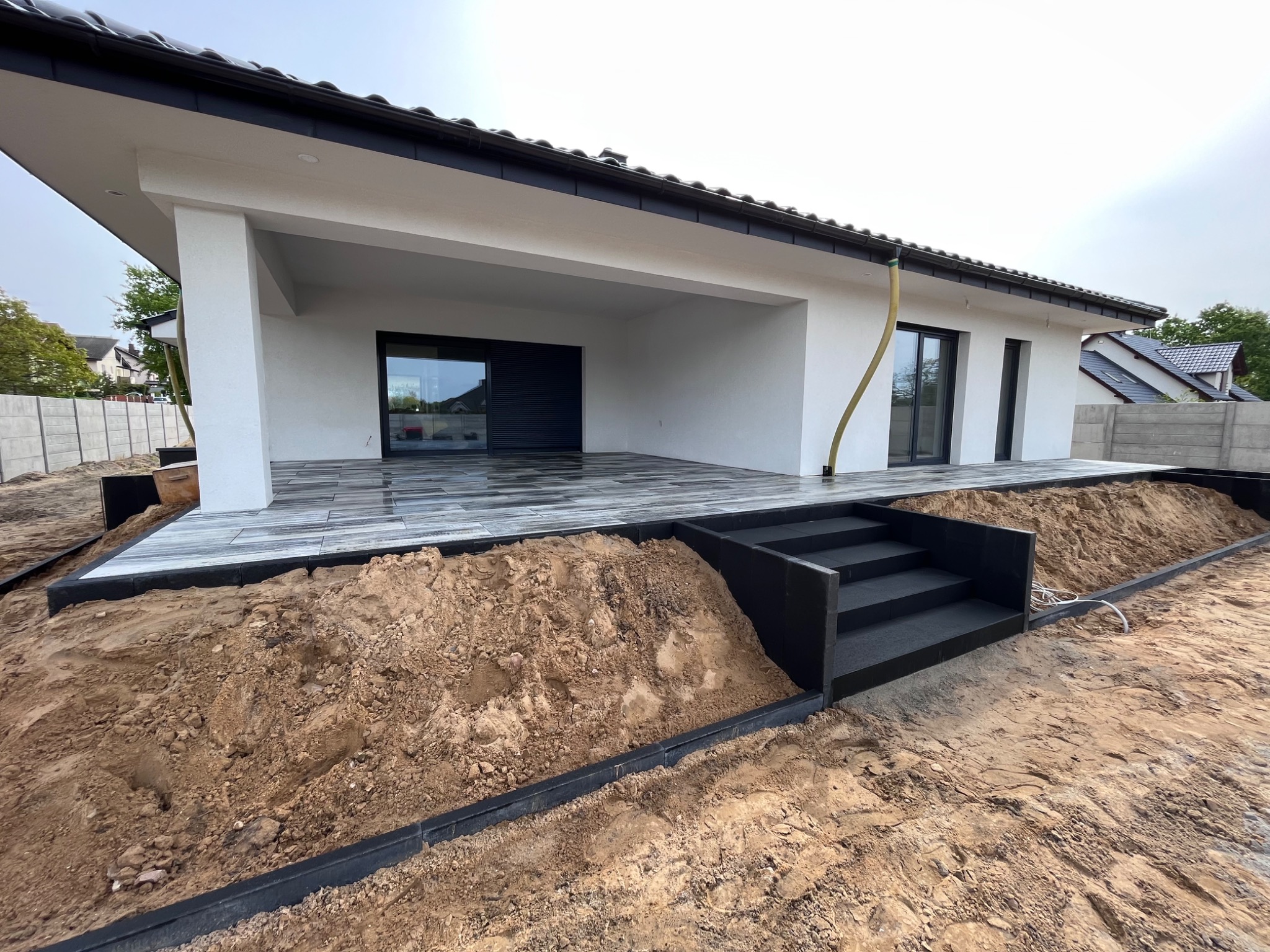 Niedokończona realizacja tarasu z kostki brukowej przy nowoczesnym domu, widoczne schody z kostki i ziemia do wyrównania terenu.