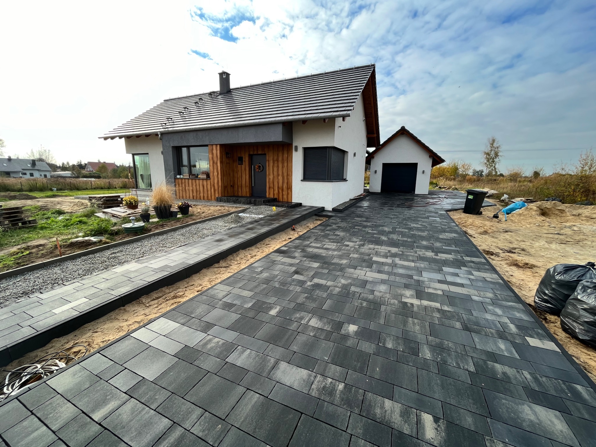 Nowoczesny dom jednorodzinny z wybrukowanym podjazdem z szarej kostki, widoczne czarne worki na śmieci i przygotowany teren pod trawnik.