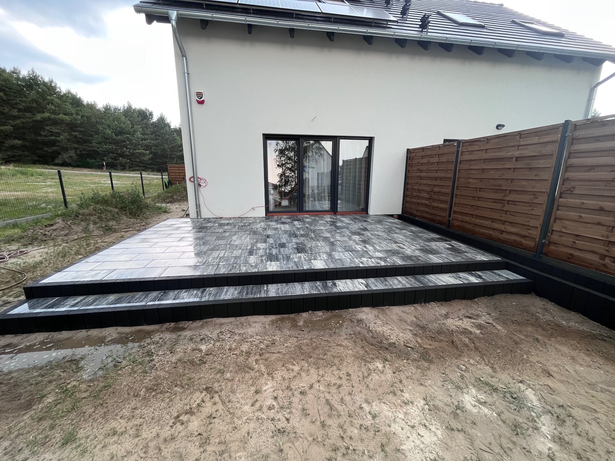 Nowoczesny taras z kostki brukowej w odcieniach szarości, z dwustopniowymi schodami, przylegający do białego domu z drewnianym ogrodzeniem w tle, widoczny fragment trawnika i drzew