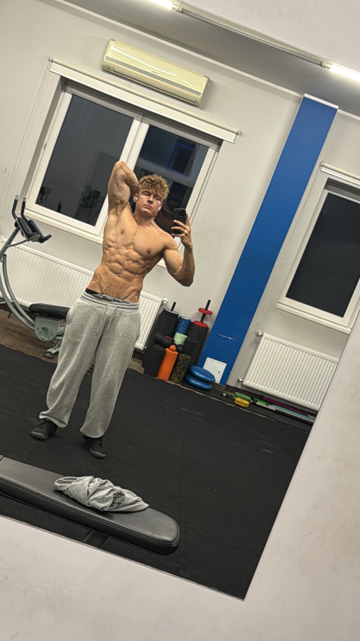 Mężczyzna z wyraźnie zarysowanymi mięśniami brzucha pozuje w szarych dresach, trzymając telefon w dłoni, na tle siłowni z oknami i sprzętem do ćwiczeń.