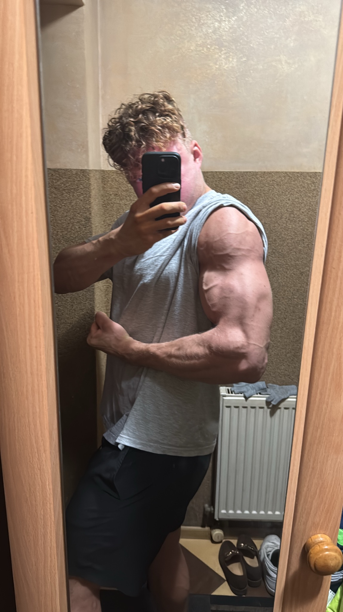 Mężczyzna w szarej koszulce prezentuje bicepsy, robiąc selfie w lustrze. Widoczne żyły na ramieniu, kręcone włosy i fragment pokoju z grzejnikiem w tle.