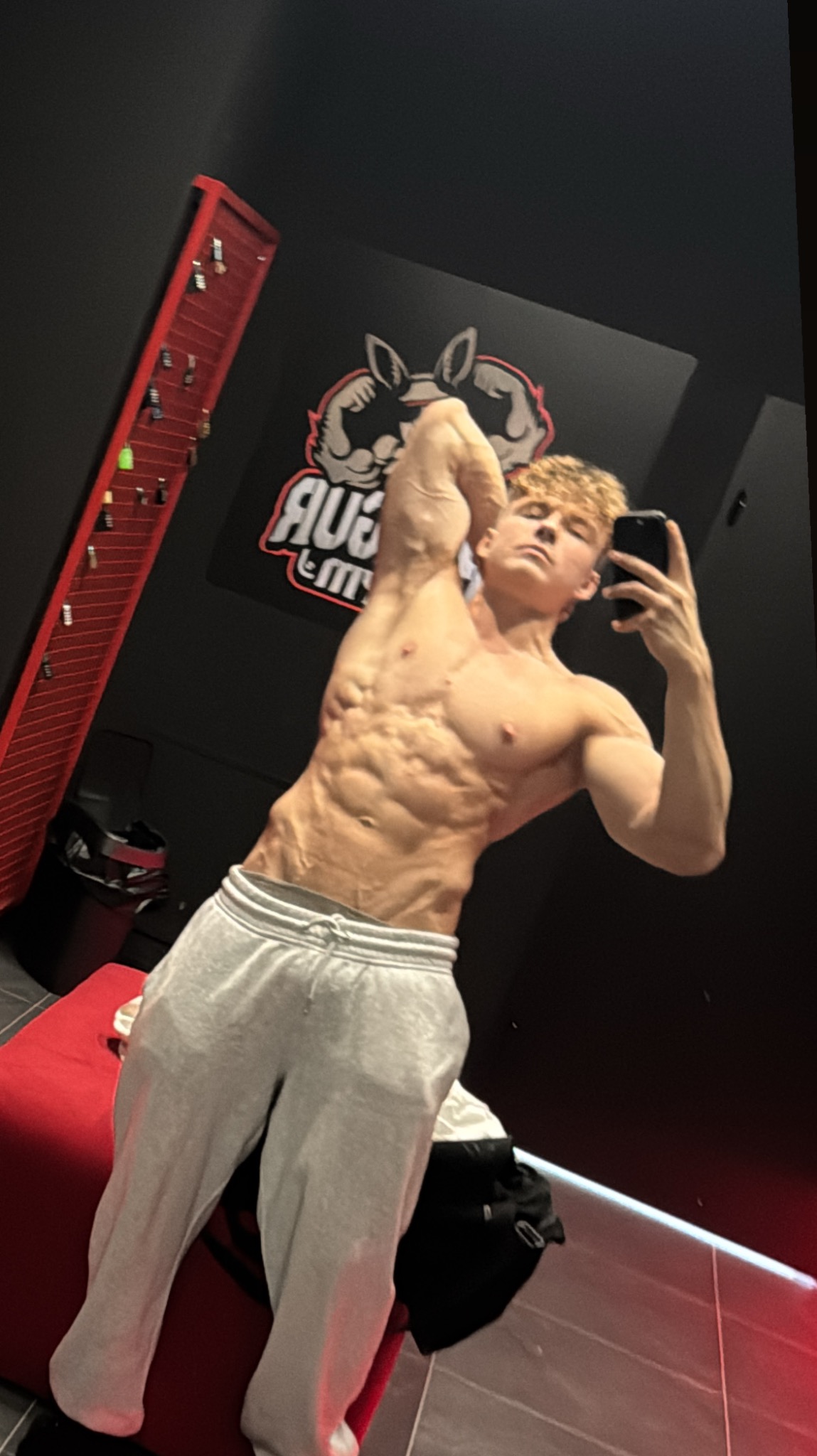 Młody mężczyzna w szarych spodniach dresowych prezentuje wyrzeźbioną sylwetkę, robiąc selfie w siłowni na tle czarnej ściany z logo. Widoczne mięśnie brzucha i ramion.