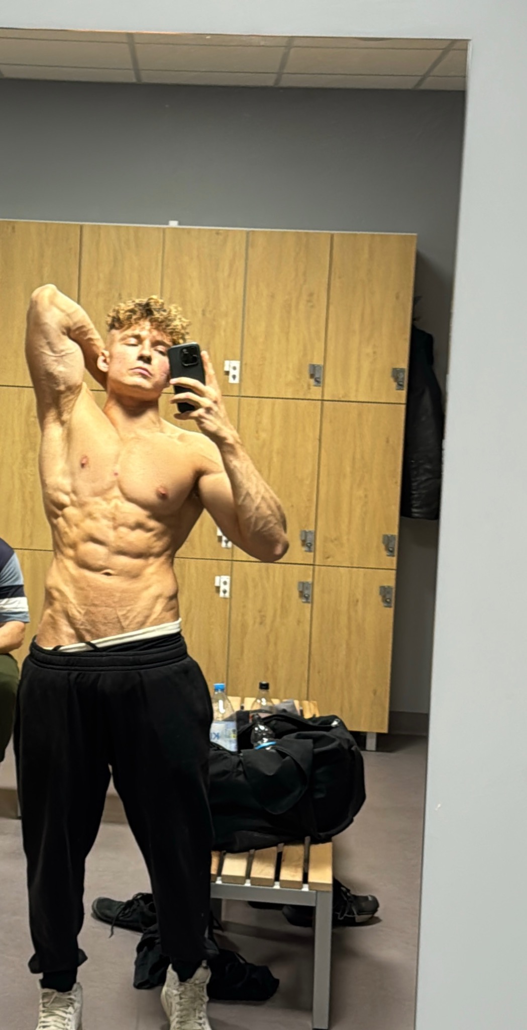 Mężczyzna z wyrzeźbionym ciałem pozuje do selfie w szatni siłowni, prezentując muskulaturę brzucha i ramion na tle szafek i torby sportowej.