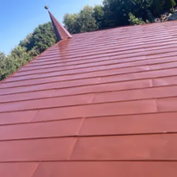 Nowe, lśniące, miedziane pokrycie dachowe z widocznym stożkowym zakończeniem, na tle zieleni drzew i błękitnego nieba.